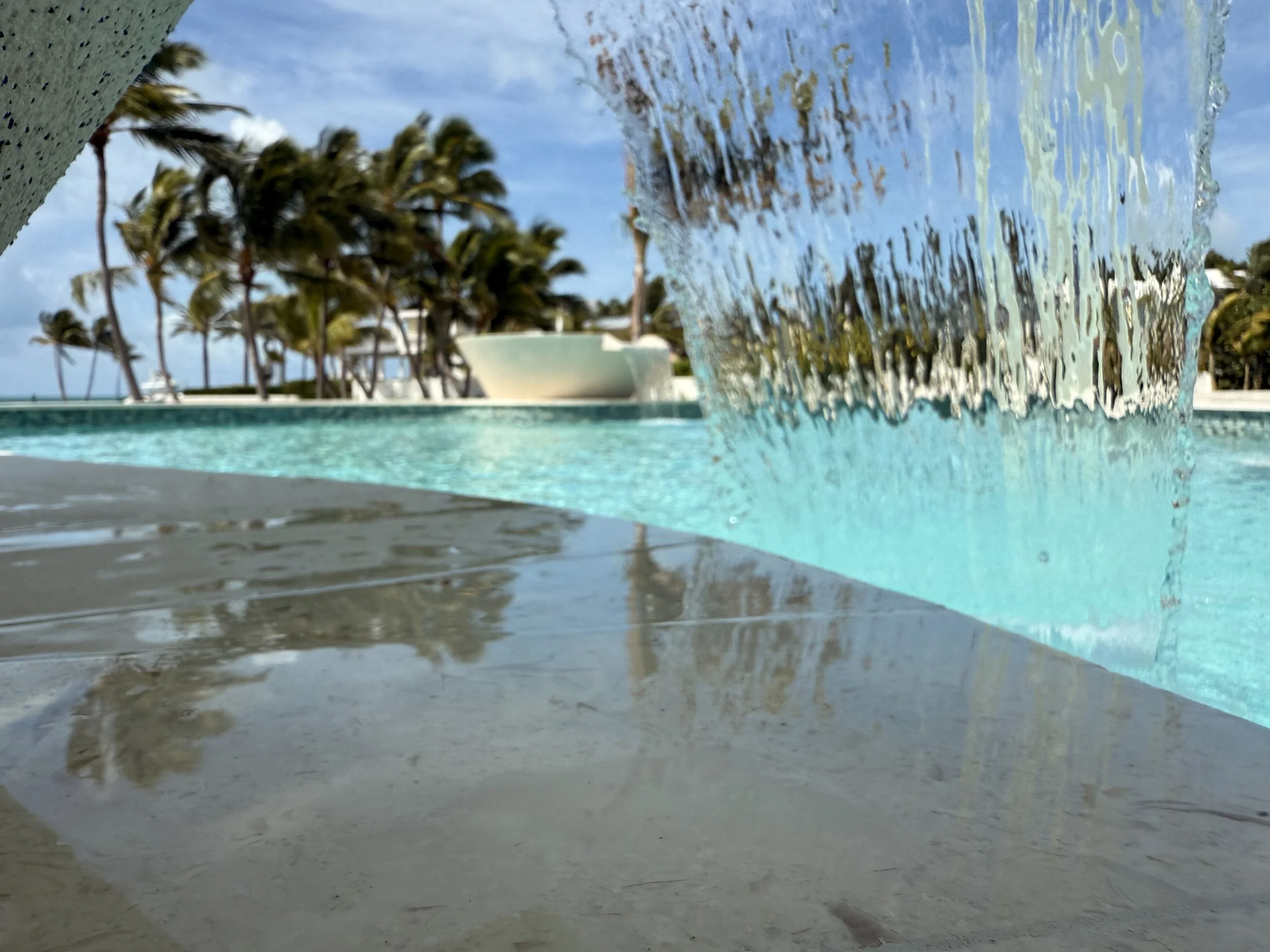 Close-up of water cascading from a waterfall feature into a pool, with palm trees and a partly cloudy sky in the background.