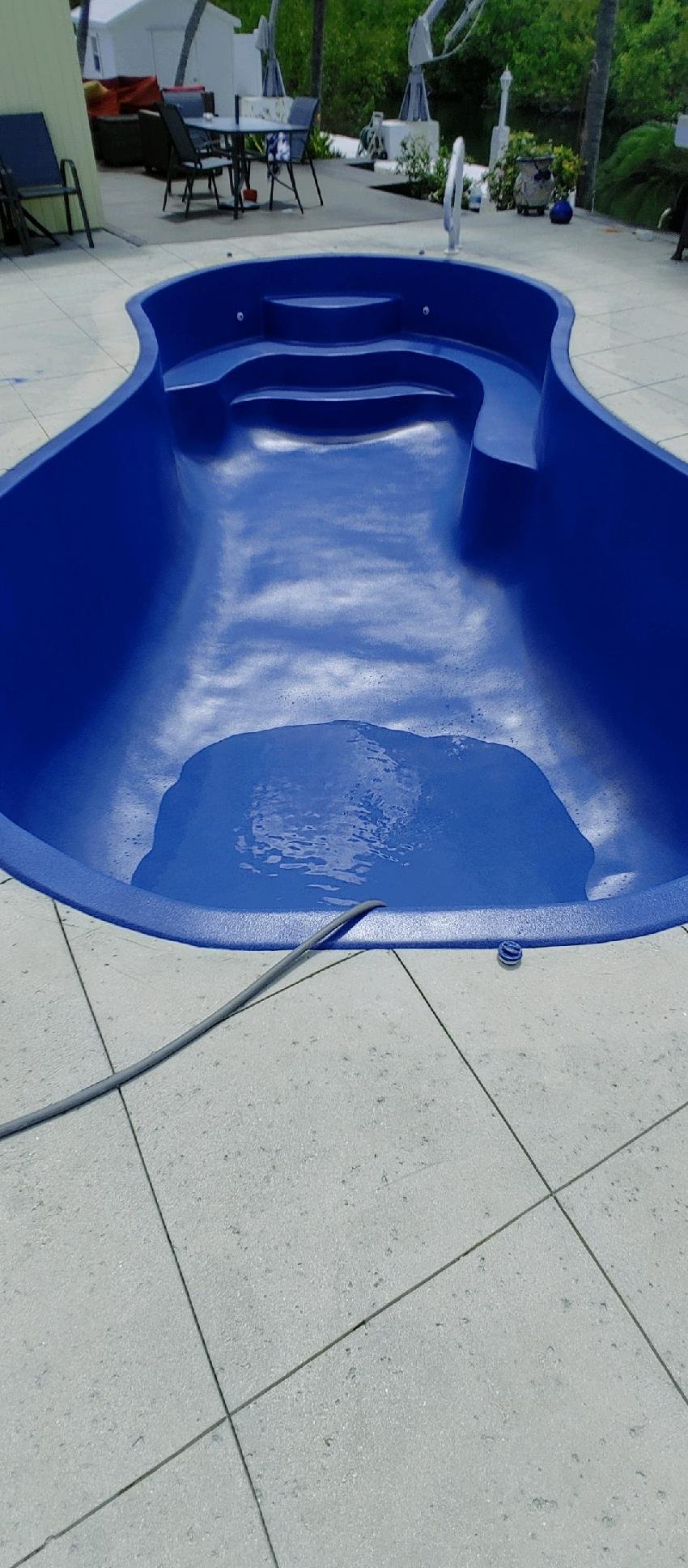 Blue kiddie pool filled with water on a concrete patio, with outdoor furniture and trees in the background.