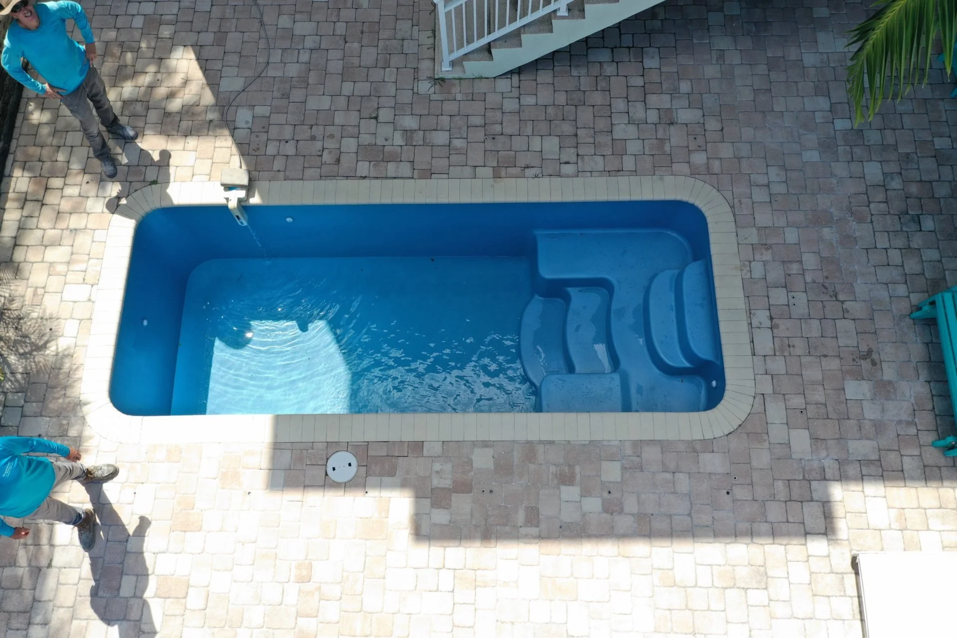 A rectangular in-ground swimming pool with blue water, surrounded by brick pavers, with steps at one end. There are two people standing near the pool, one on the left and one at the top, and a woman sitting on a turquoise bench on the right. A palm tree in the top right corner casts part of its shadow on the ground.