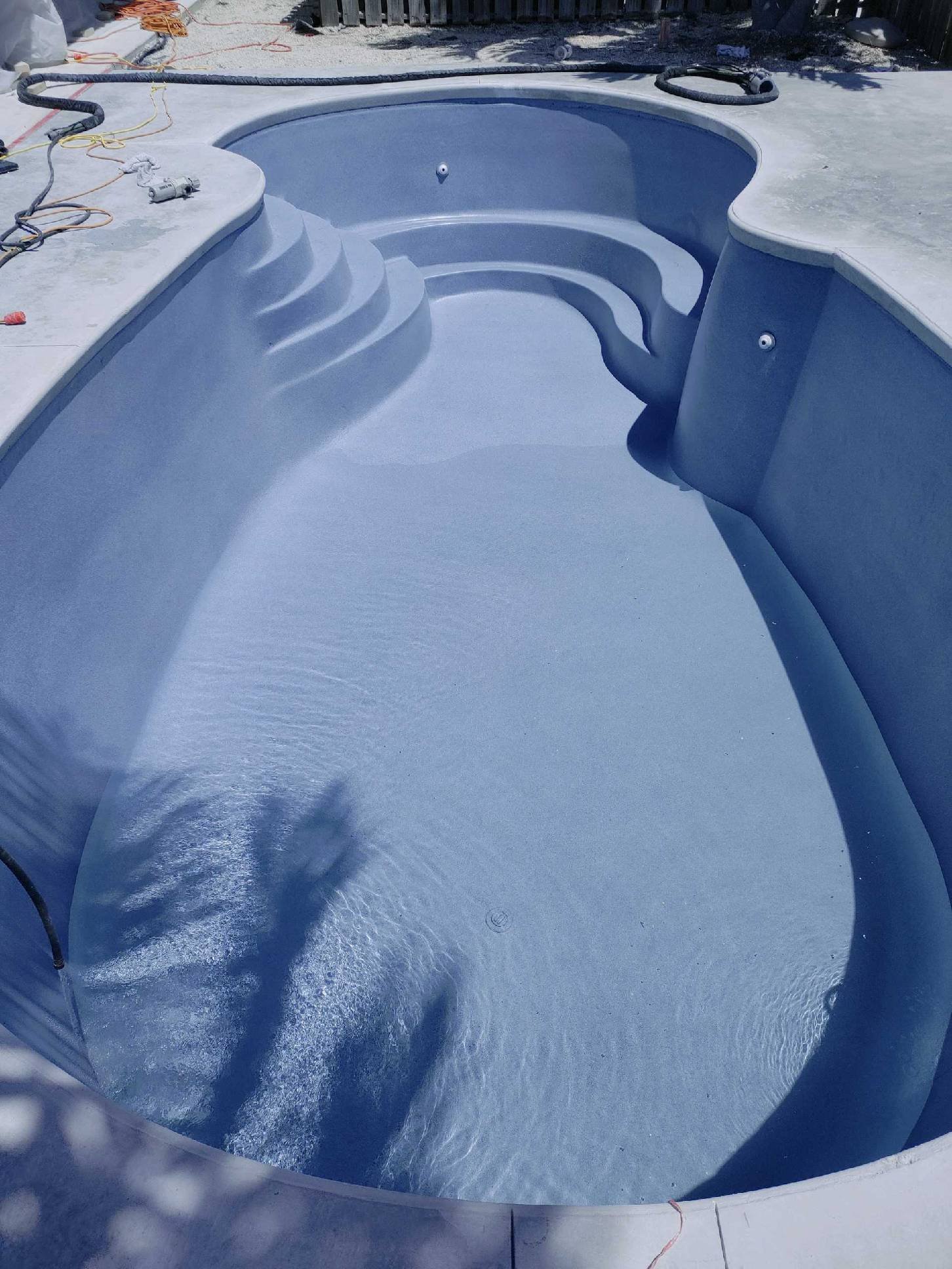 Empty in-ground swimming pool under construction with stairs inside and construction tools around the edge.