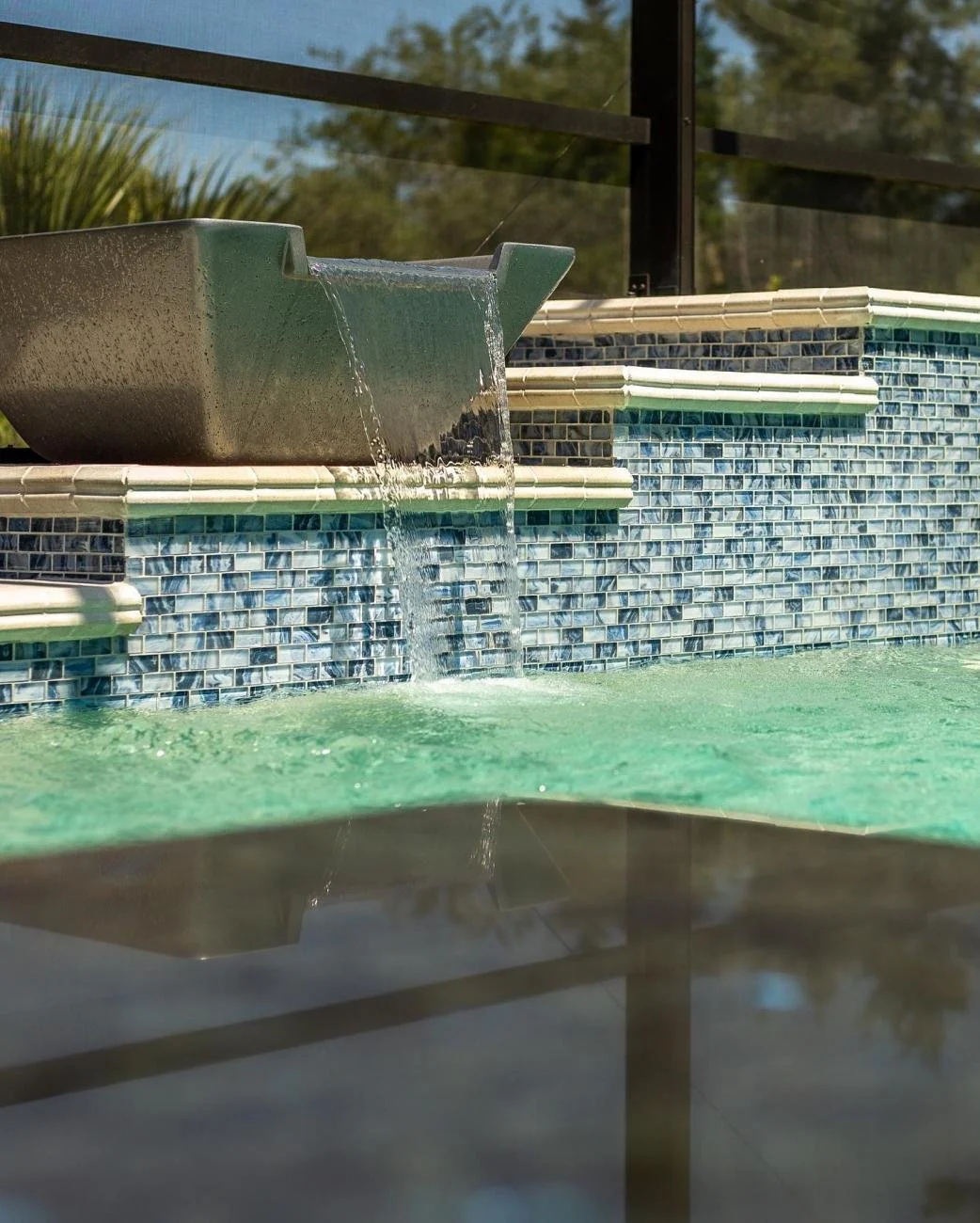 Close-up of a fountain spout with water cascading into a swimming pool, tiled wall in the background, sunlight reflecting on the water.