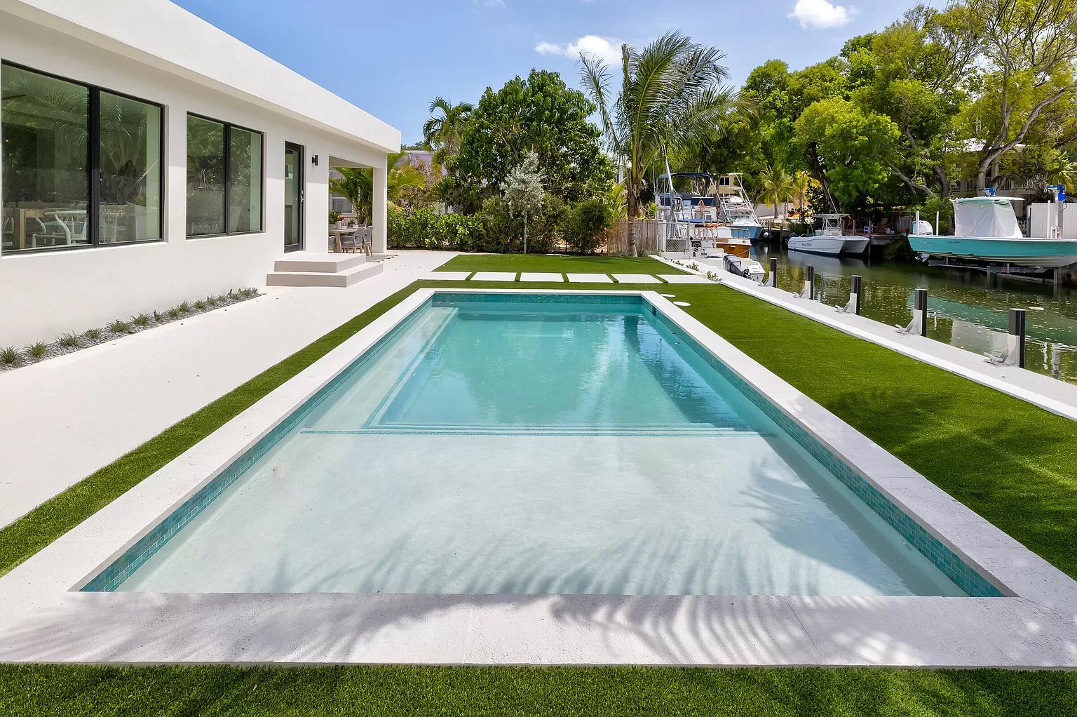 Modern backyard with a rectangular in-ground pool, green artificial grass accents, white concrete decking, and boat marina with boats docked along waterway, surrounded by lush trees and a white contemporary house.