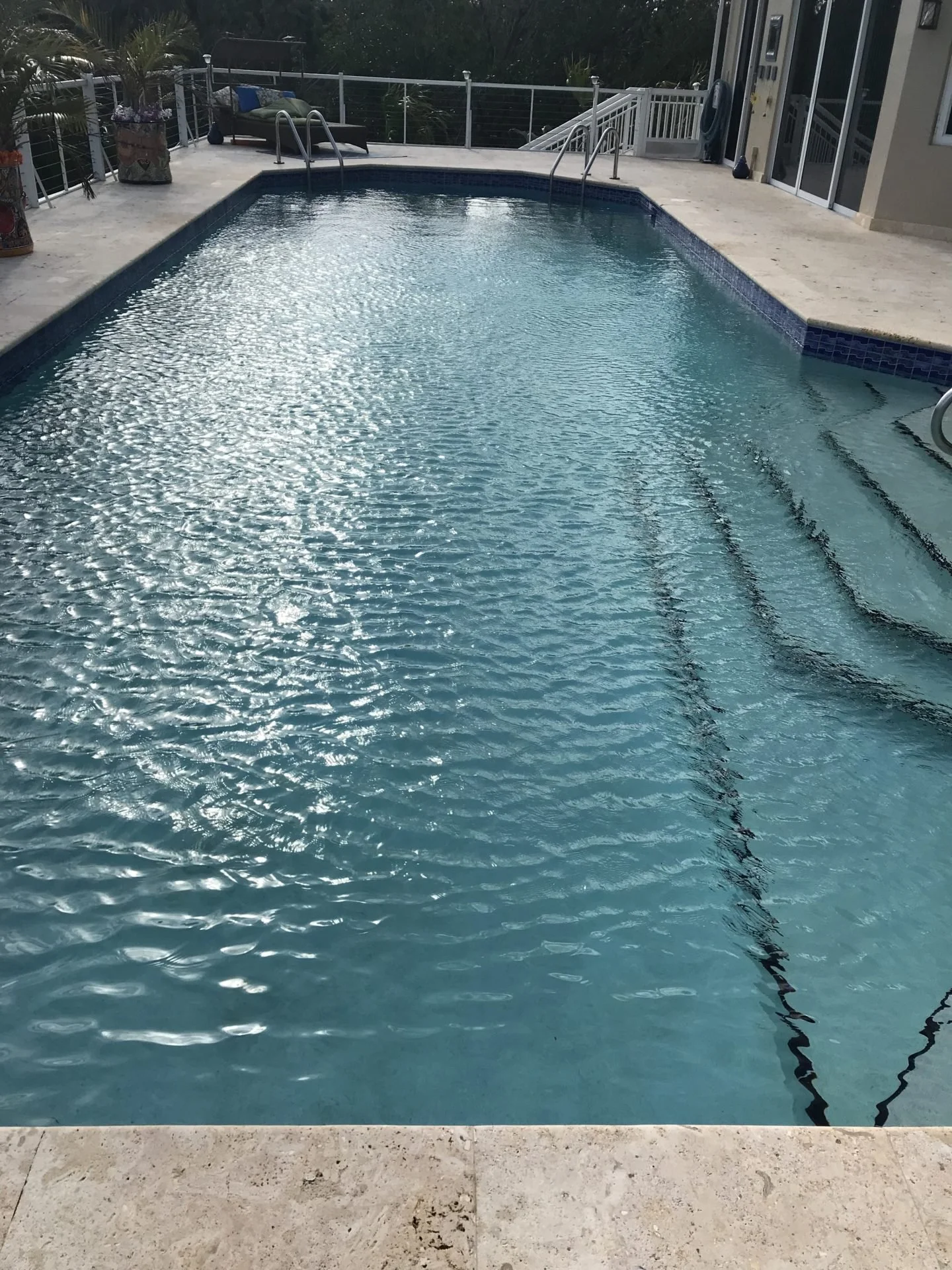 Residential backyard swimming pool with stairs, surrounded by a concrete deck, with a lounge chair, several potted plants, and a house exterior visible in the background.