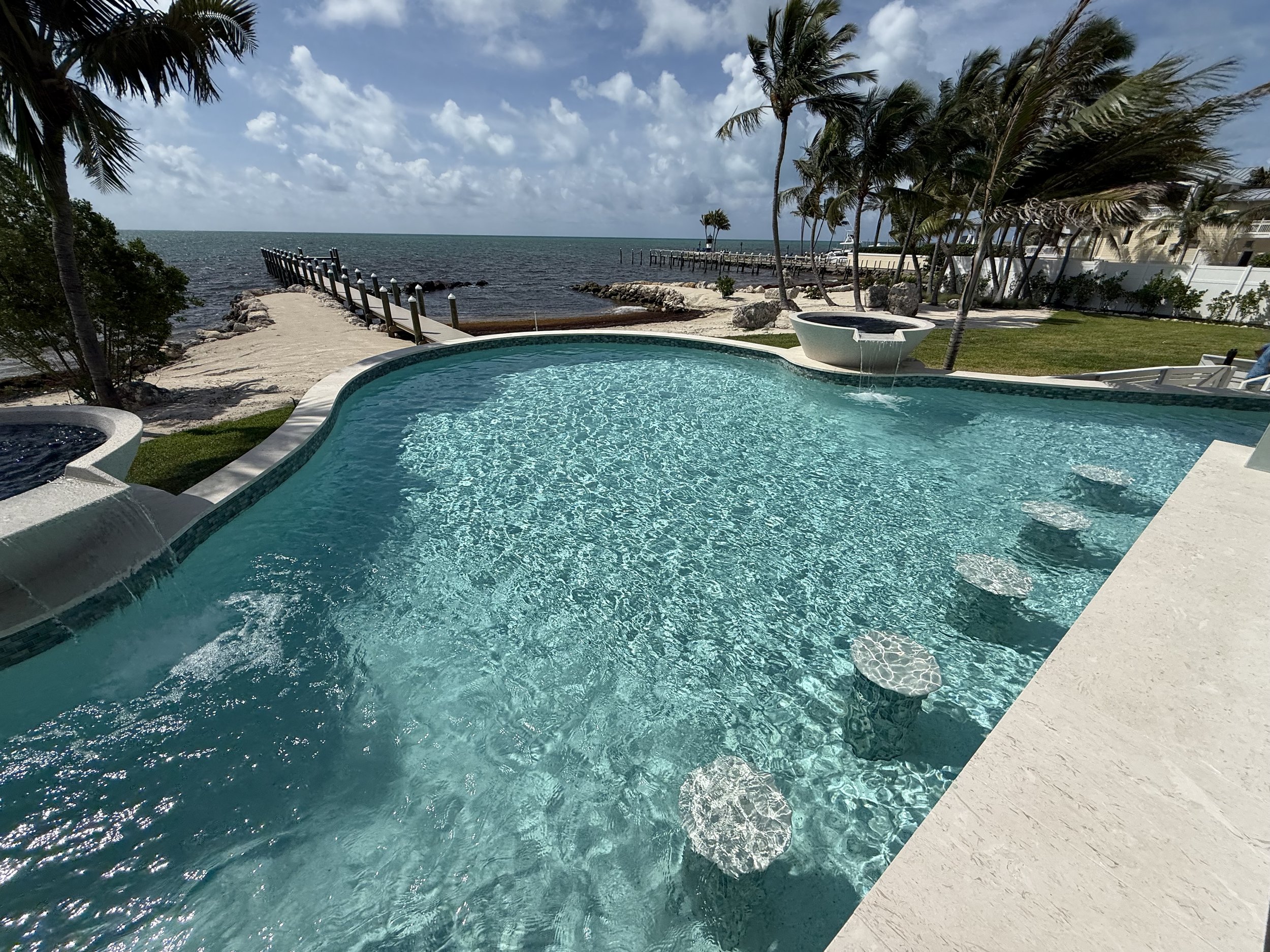 Infinity pool overlooking a beach with a pier, surrounded by palm trees and a partly cloudy sky.