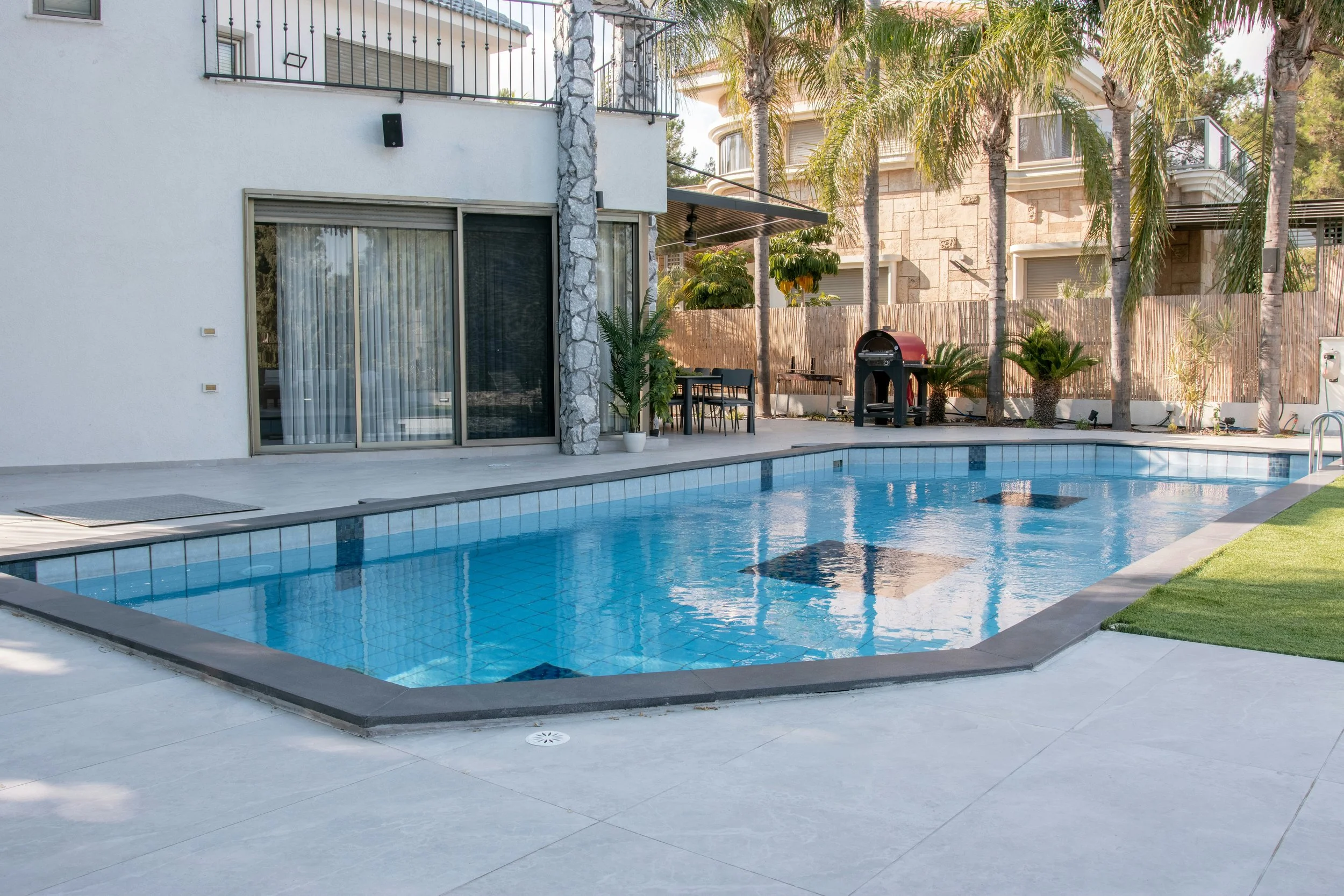 Residential backyard with a clear blue swimming pool, surrounded by a concrete deck, potted plants, palm trees, and a wooden privacy fence. A house with large sliding doors, a dining table with chairs, and a barbecue grill are visible in the backgrou