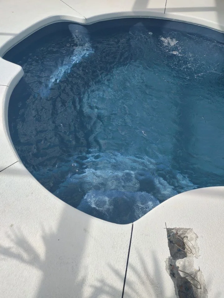 A small, round hot tub with clear blue water outdoors, surrounded by a white concrete deck, with a small hole in the deck near the bottom right corner.