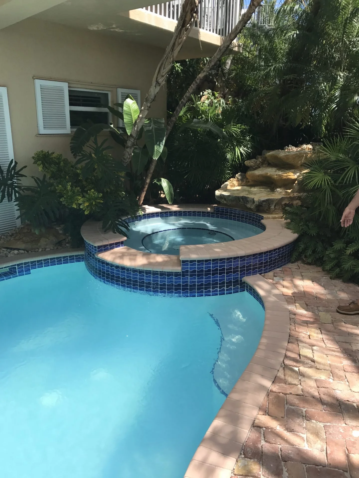 Residential backyard pool area with a swimming pool and a hot tub surrounded by tropical plants and stone features.