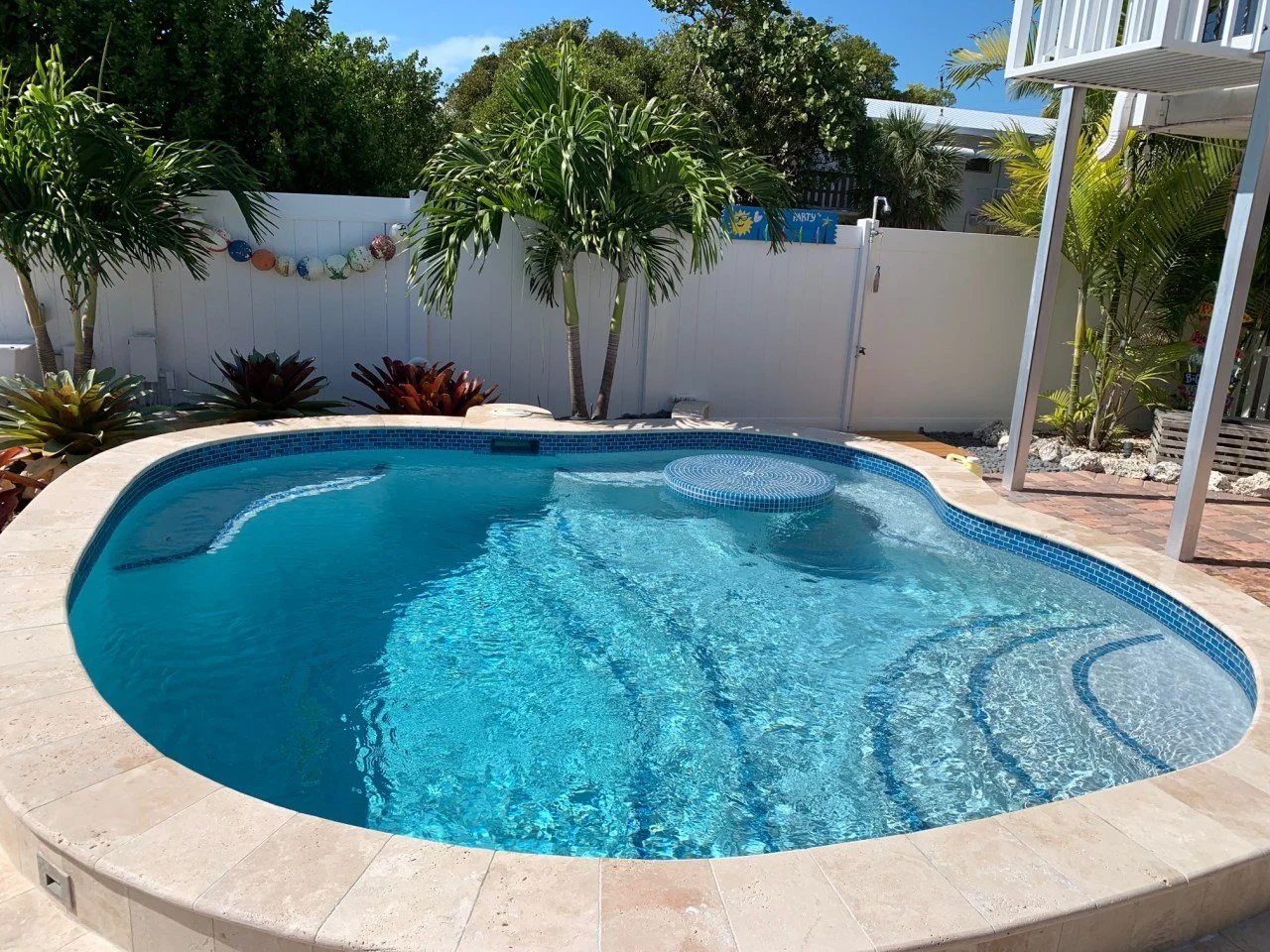 A backyard swimming pool surrounded by a stone deck, with tropical plants and trees along a white fence, under a clear blue sky.