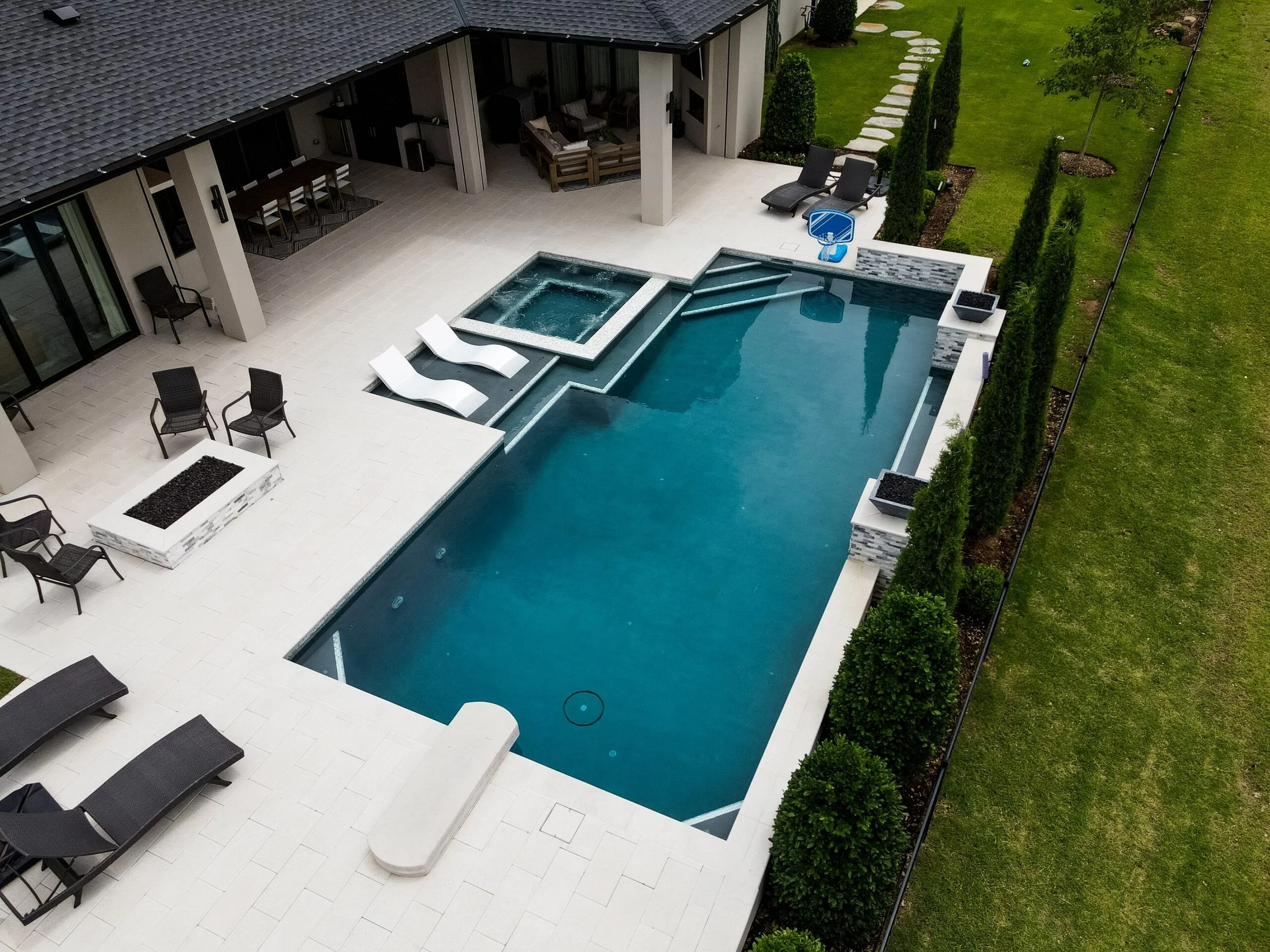 A backyard swimming pool area with a hot tub, lounge chairs, and patio furniture surrounded by a well-maintained lawn and tall bushes.