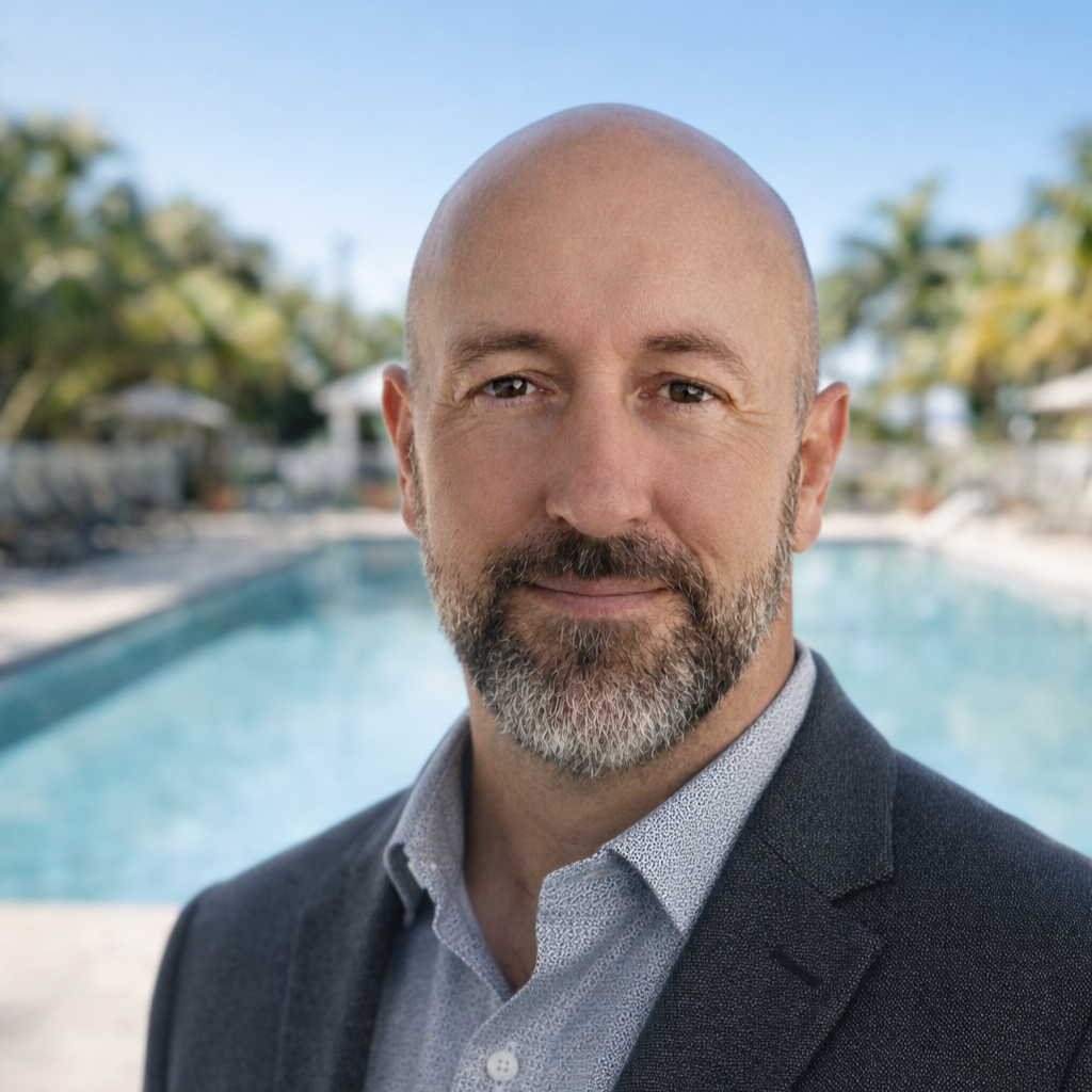 A smiling man in a blue suit and light blue shirt stands in front of a swimming pool surrounded by palm trees and lounge chairs on a sunny day.