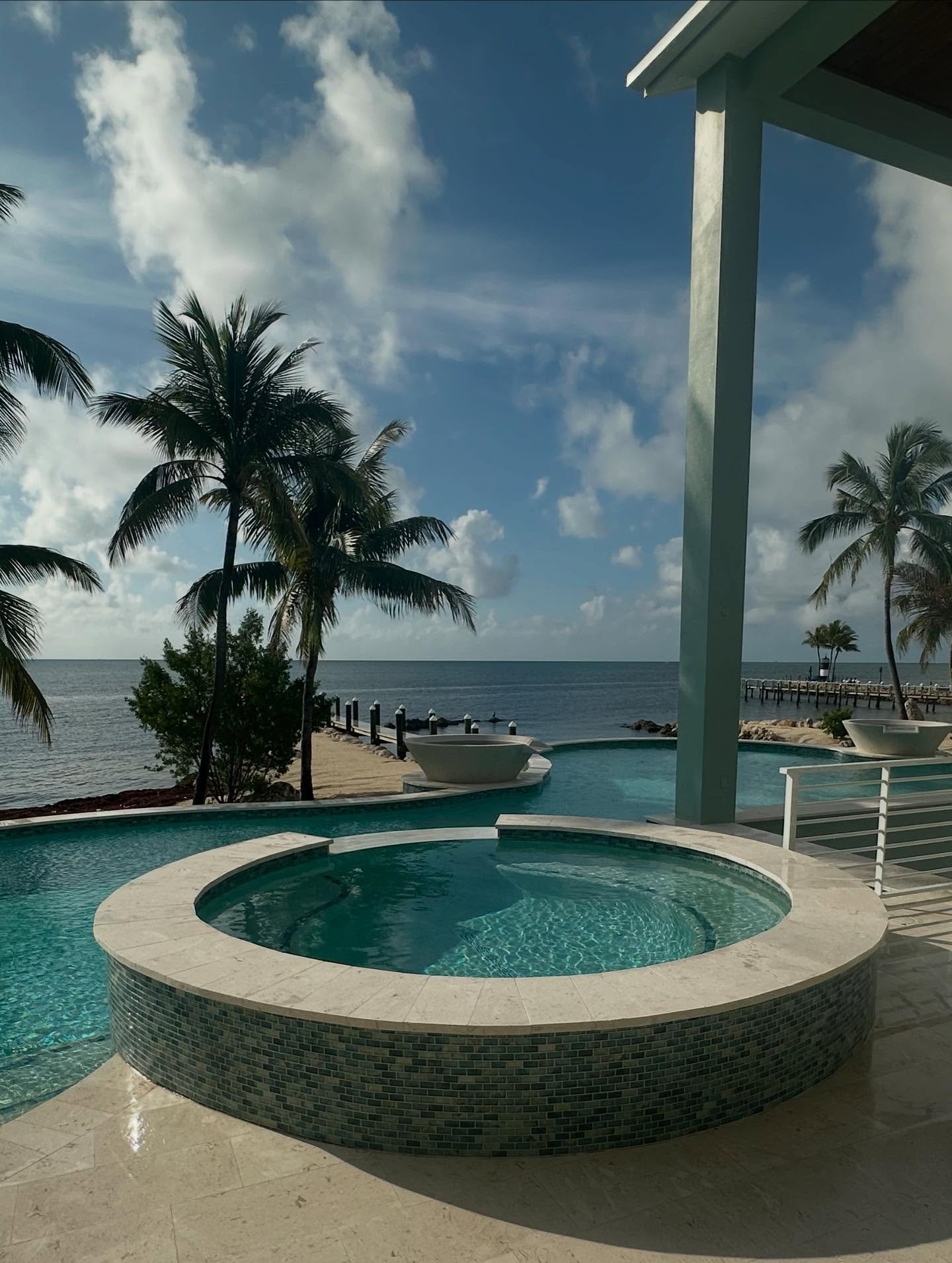 A view of a tropical resort featuring a circular hot tub with mosaic tile edges, an infinity pool, palm trees, and the ocean in the background under a partly cloudy sky.
