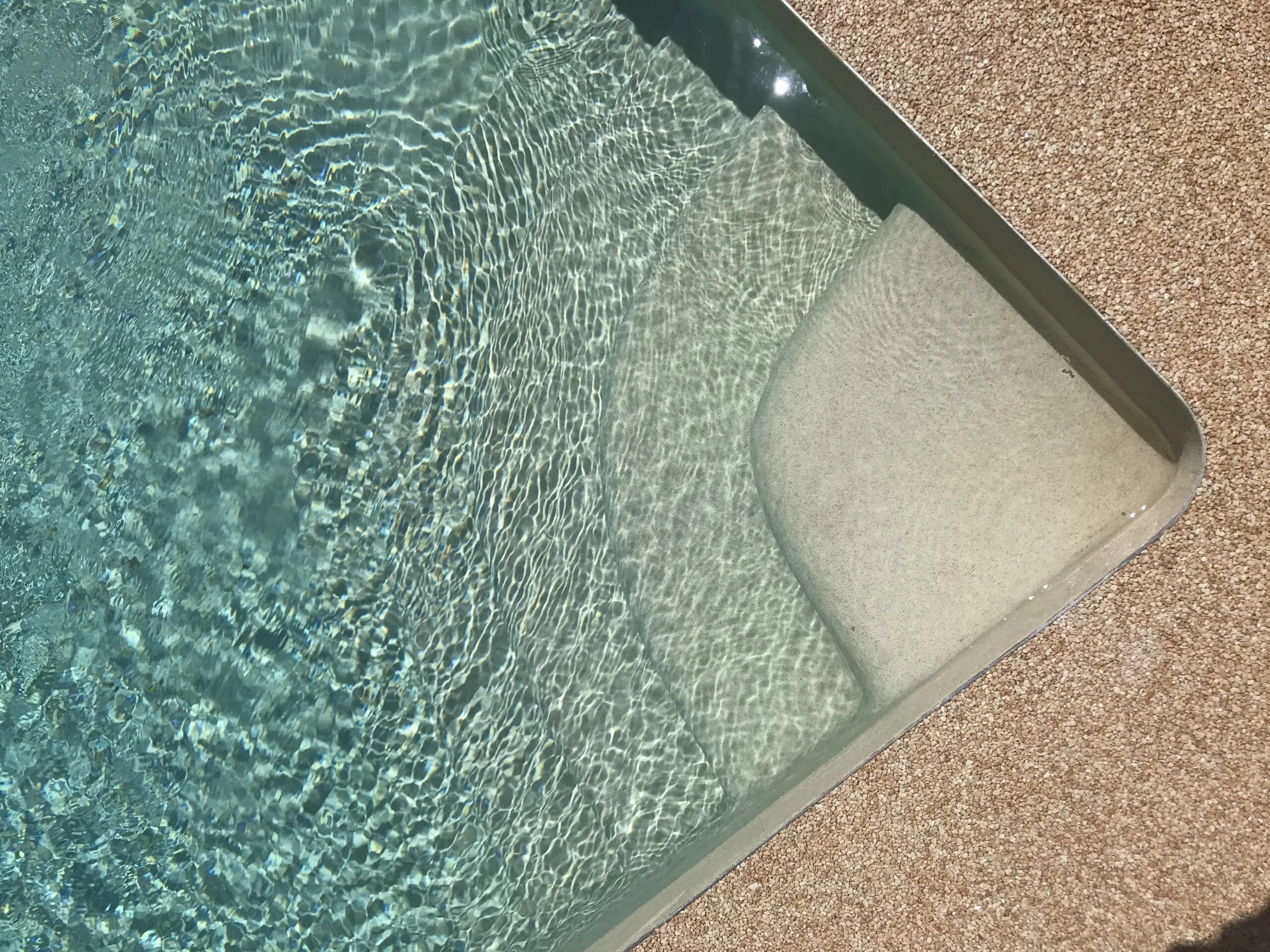 Close-up of a swimming pool with clear water, a submerged step, and textured beige poolside surface.