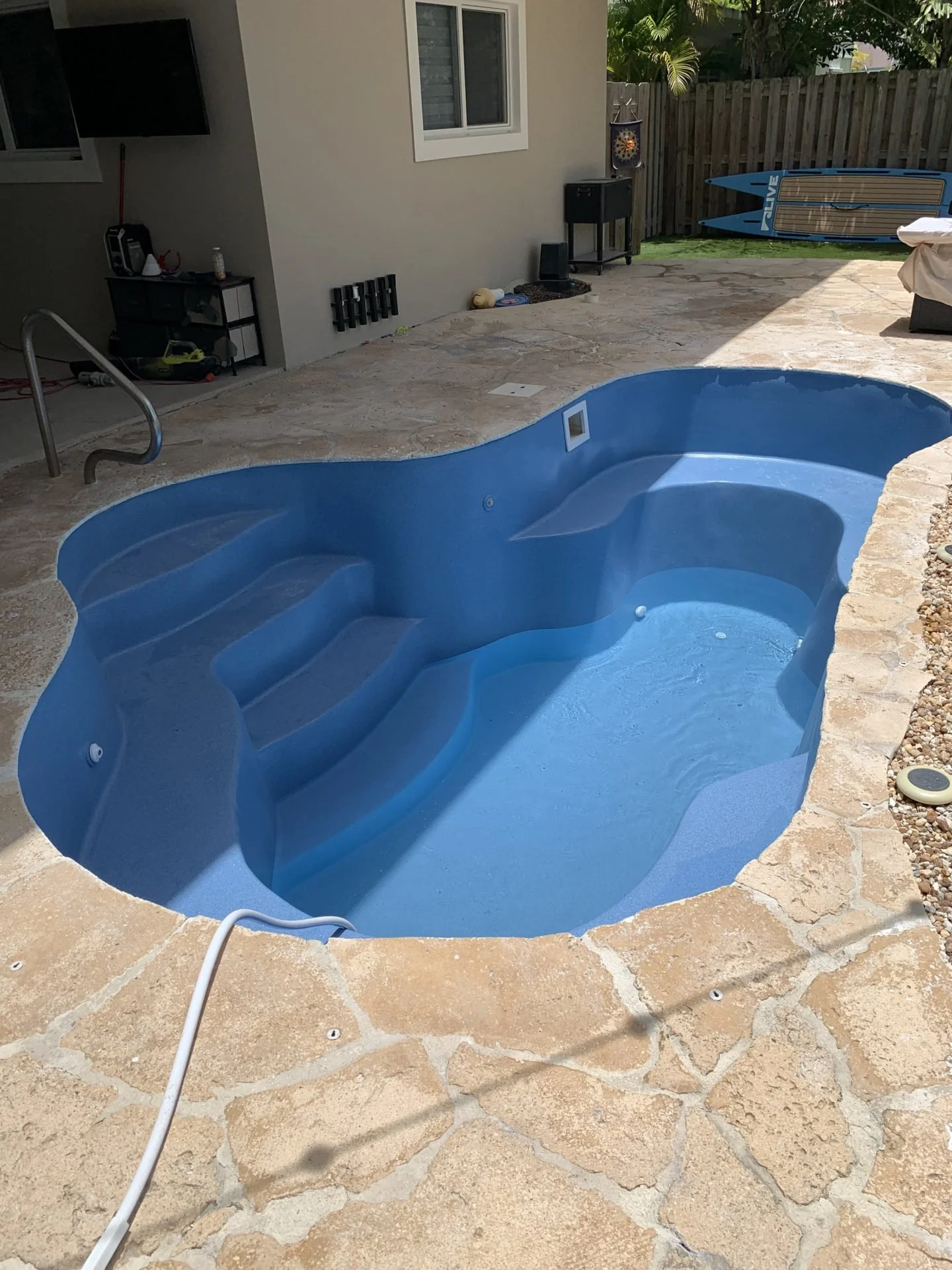 Empty backyard swimming pool with built-in steps, surrounded by stone patio, with a house wall, window, and gardening tools in the background.