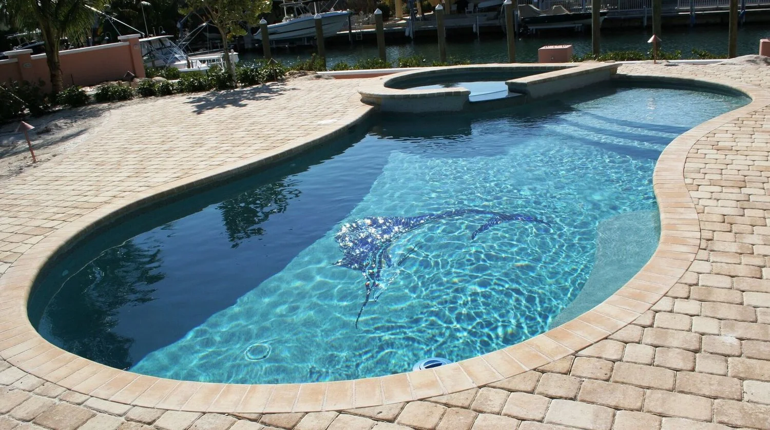 A backyard swimming pool with a rounded shape, surrounded by a brick patio. There is a built-in hot tub attached to the pool, and a mural of a dolphin is visible underwater at the bottom of the pool. In the background, there are boats docked at a mar