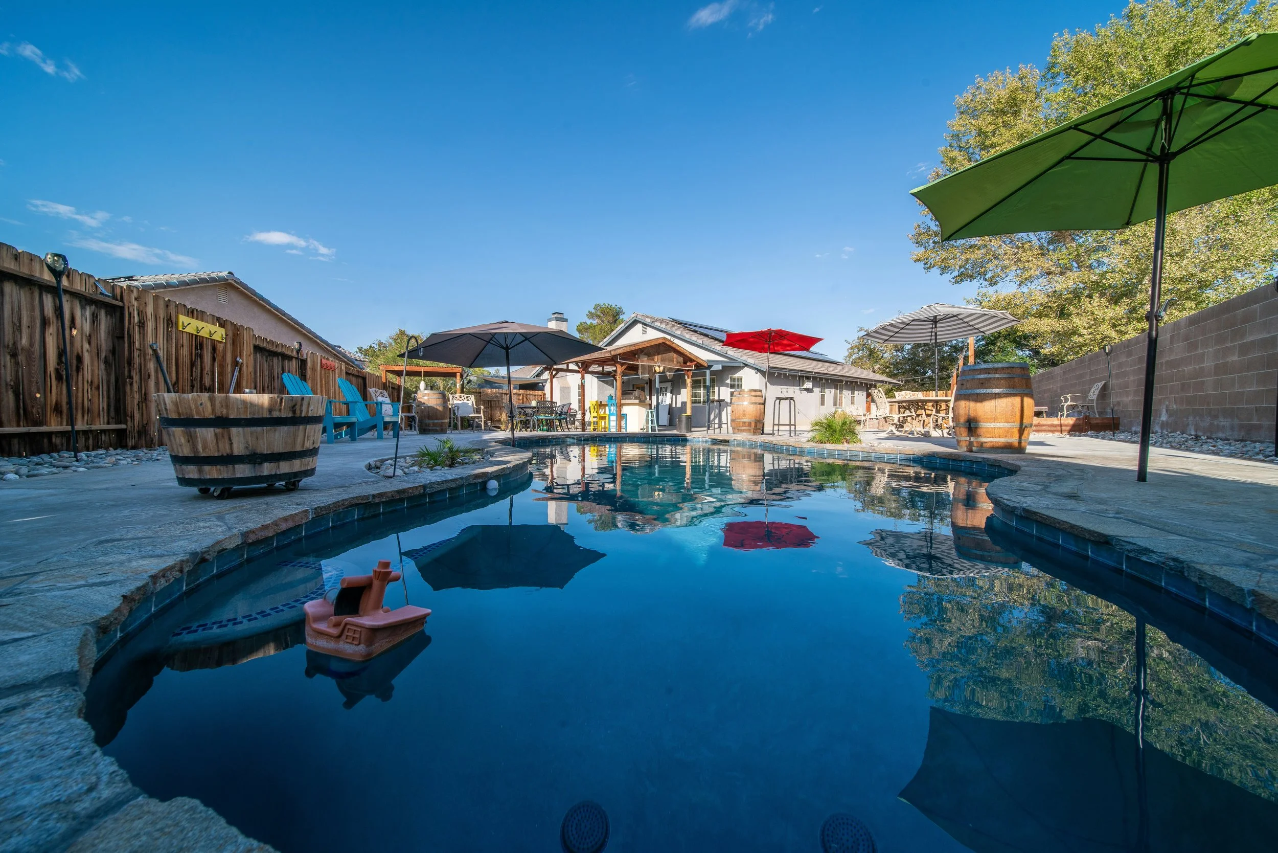 Backyard swimming pool with umbrellas, lounge chairs, and a shaded sitting area, surrounded by a fence and trees.