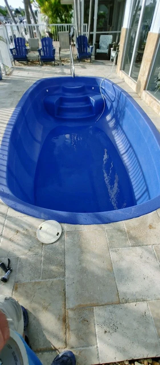 Empty blue plastic hot tub on tiled outdoor patio with pool chairs and glass doors in background.