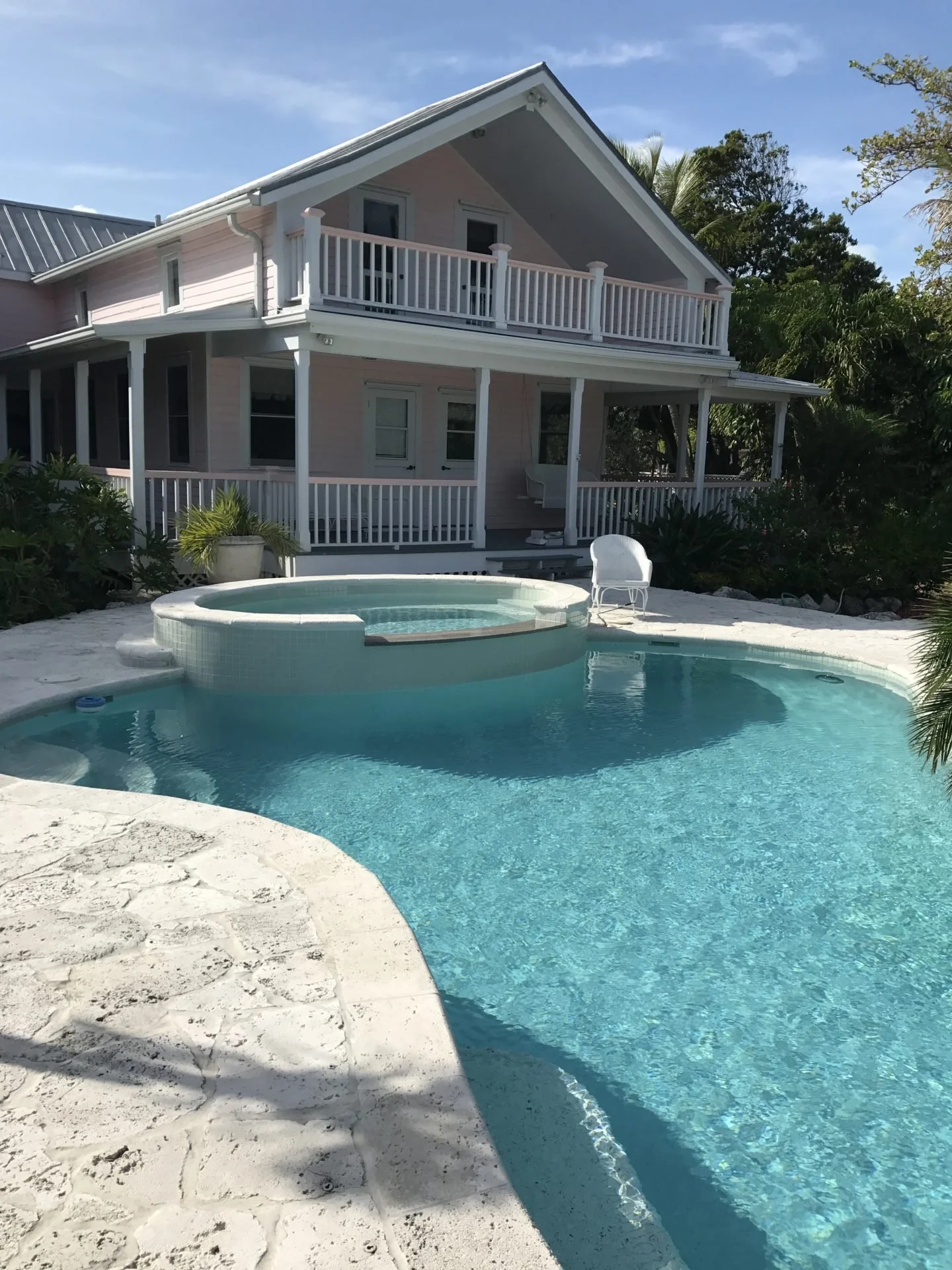 A pink two-story house with white railings and a covered porch, sitting behind a kidney-shaped swimming pool with a raised hot tub, surrounded by a white stone deck, with lush greenery and trees in the background.