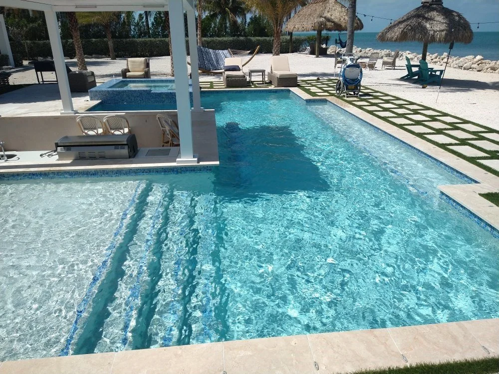 A swimming pool area with steps, lounge chairs, umbrellas, and a view of the beach and ocean in the background.