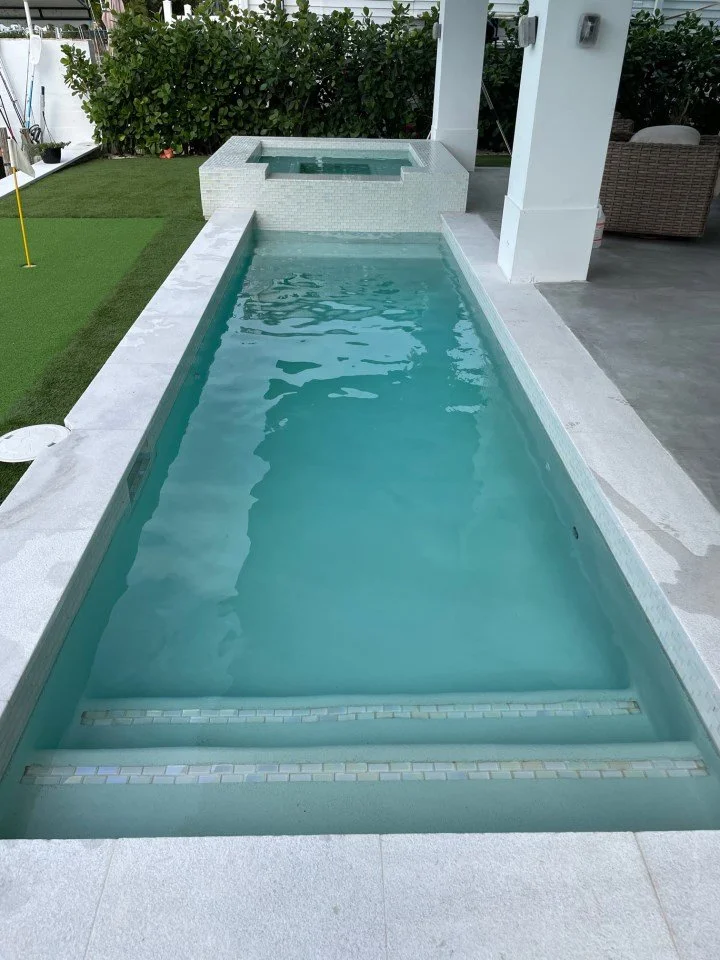 An outdoor backyard pool with steps leading into the water, with a hot tub in the background, surrounded by manicured grass and some outdoor furniture.
