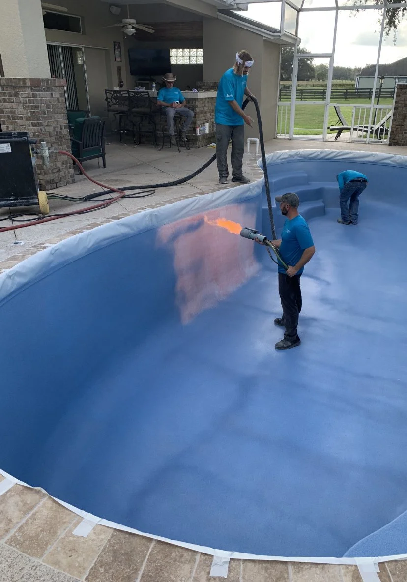 Workers are cleaning and preparing an in-ground swimming pool using a heat gun, with one person operating the heat gun and others assisting inside the pool. Two individuals are resting on the patio area in the background.
