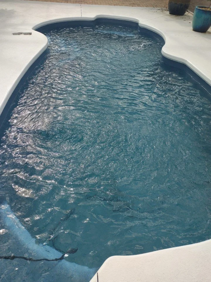 A swimming pool outdoors with blue water, surrounded by a white concrete deck and potted plants.