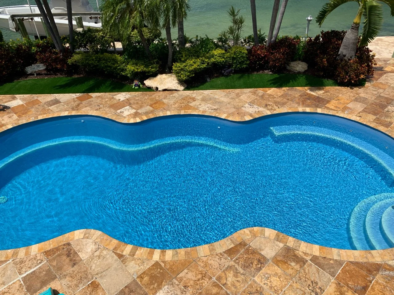 A blue, kidney-shaped swimming pool with stone tile deck, greenery, small palm trees, and water view beyond.