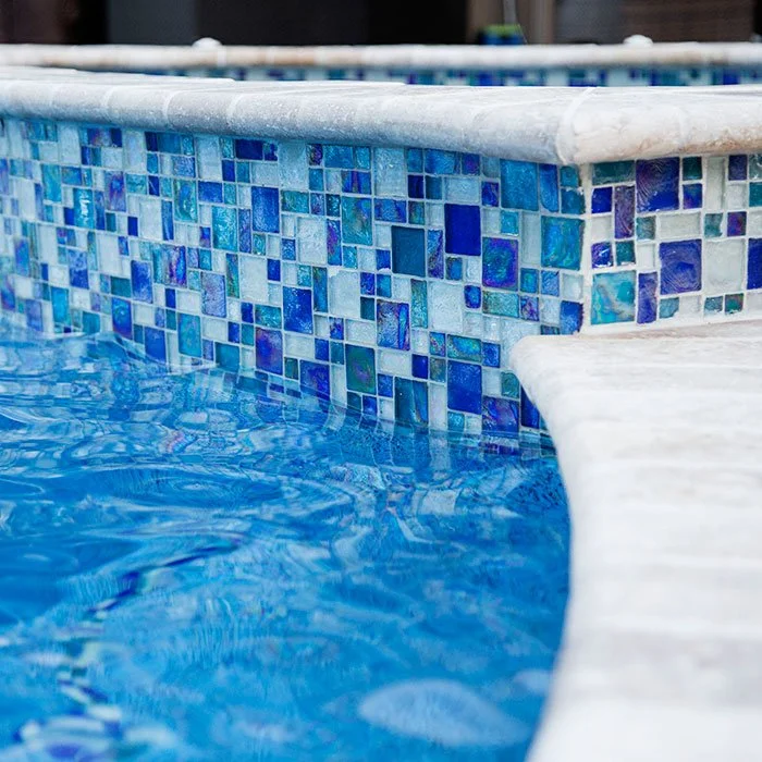 Close-up of a swimming pool edge with blue mosaic tile border and water reflected underneath.