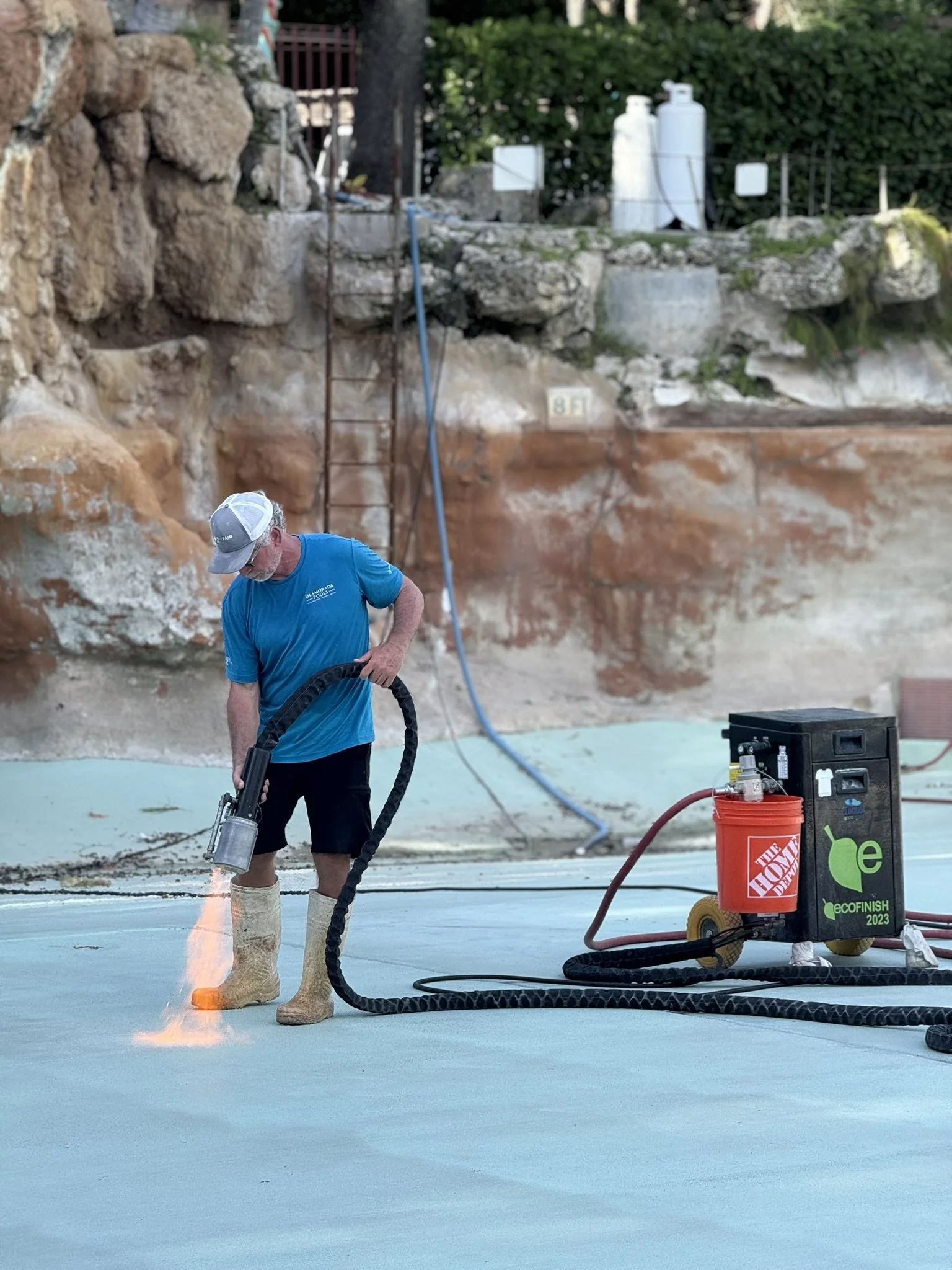 A man working on a concrete surface, using a high-pressure torch to smooth or treat it, with equipment labeled EcoFinish and Home Depot nearby.