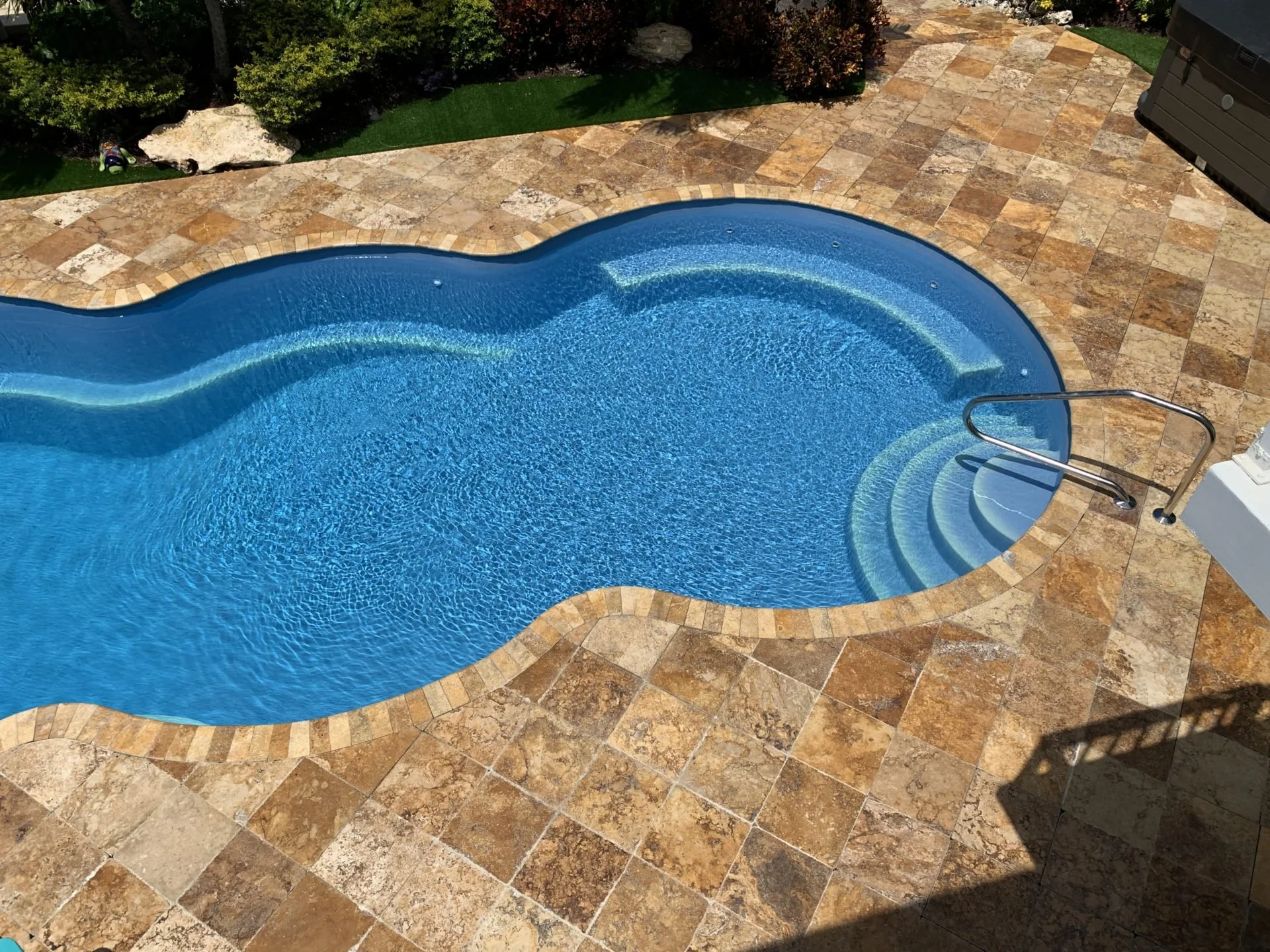 An outdoor swimming pool with curved steps, surrounded by a stone deck and landscaped greenery.