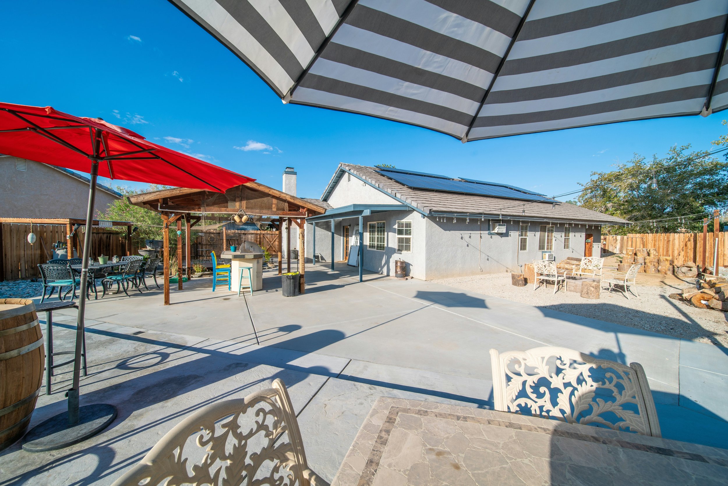 Backyard patio with tables, umbrellas, a barbecue area, seating, and a house with solar panels on the roof.