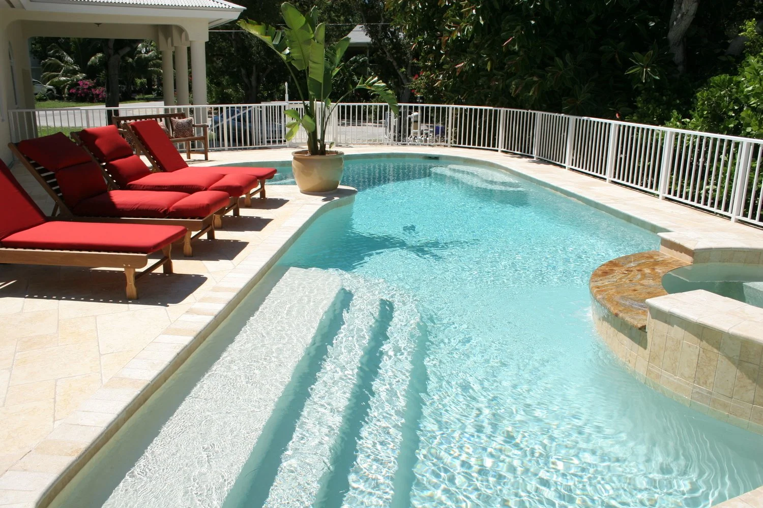 Vacation pool with red lounge chairs, potted plant, and lush greenery in the background.
