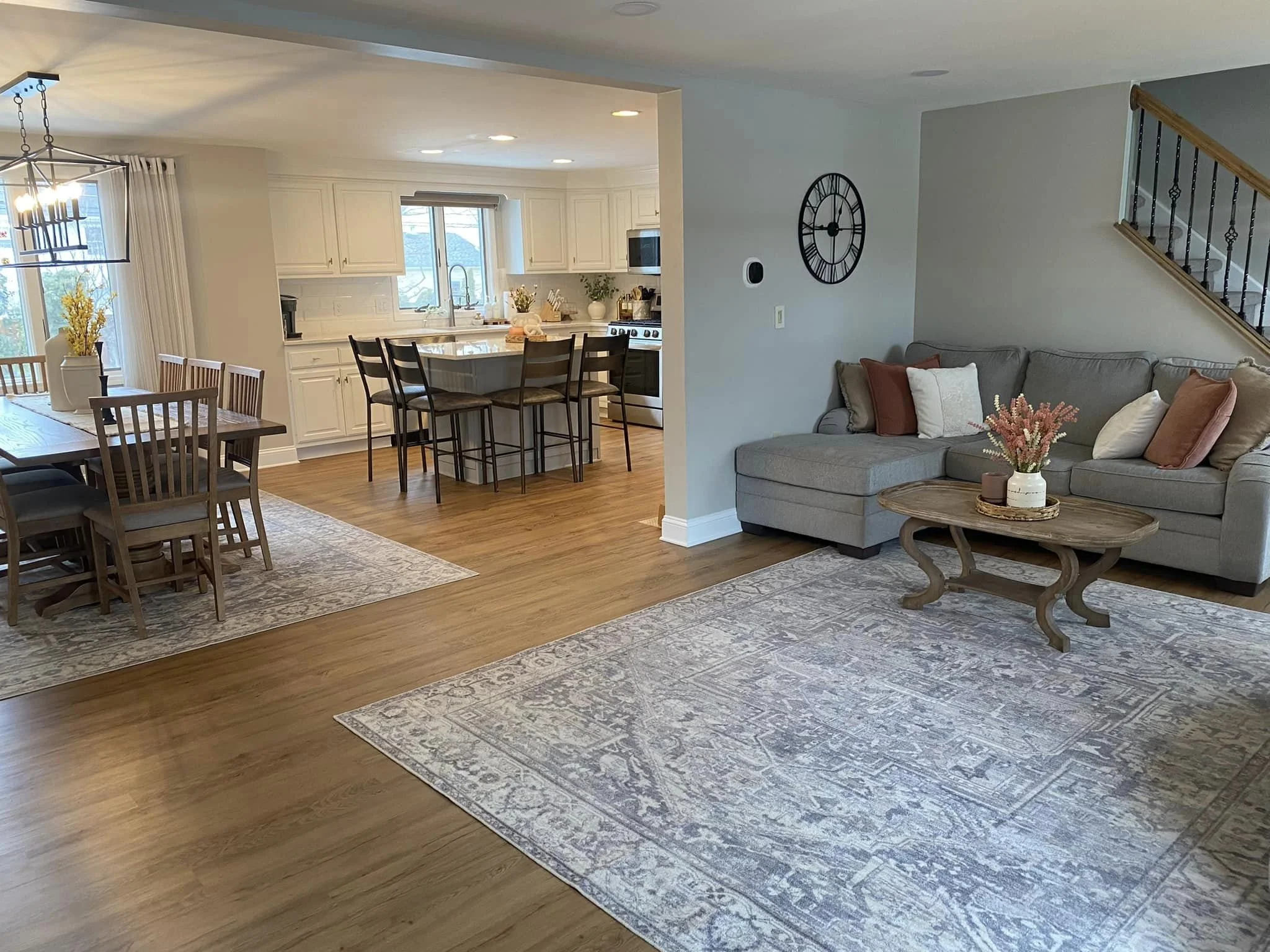 Open-concept living room and kitchen with wooden flooring, a gray sofa with cushions, a coffee table, a dining table with chairs, and a kitchen with white cabinets and a breakfast bar.