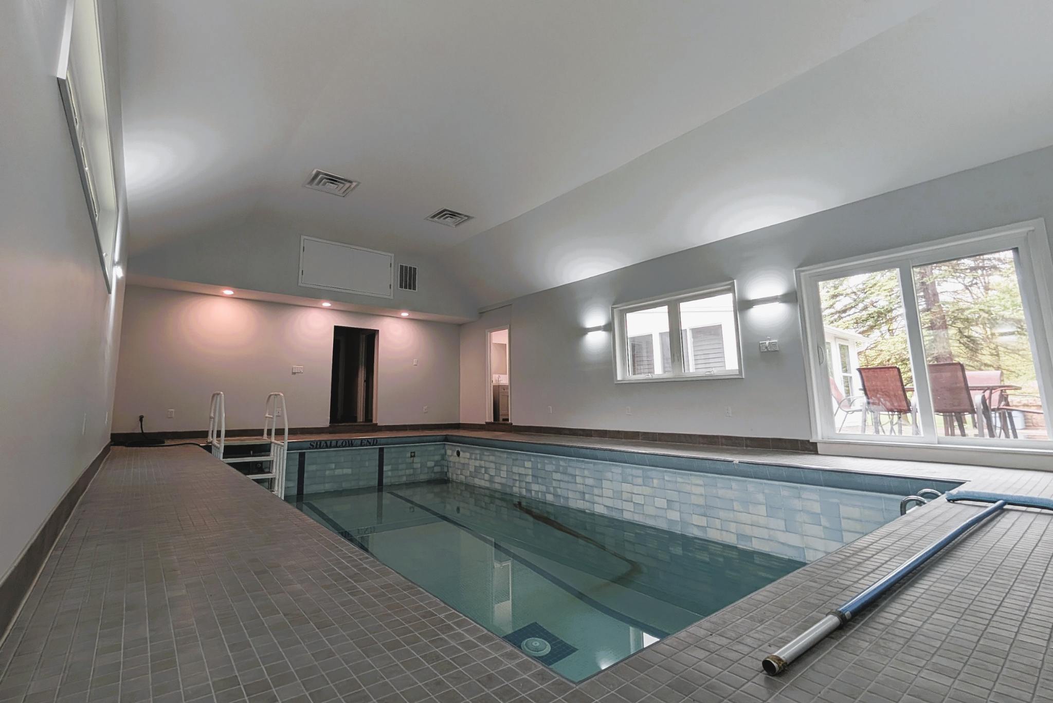 Indoor swimming pool with beige tiled floor, white walls, and large windows with an outdoor patio visible through sliding glass doors.