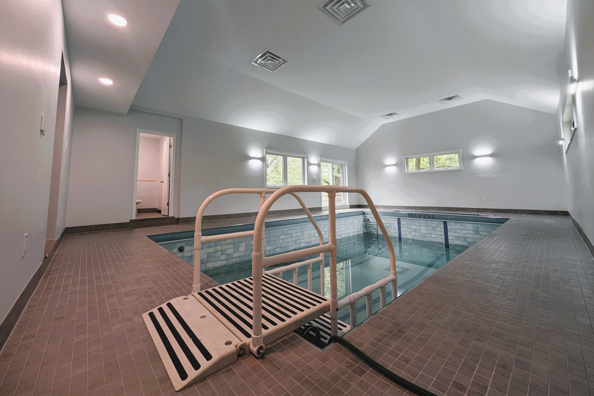 Indoor swimming pool with a white pool lift, tiled floor, white walls, several windows, and ceiling lights.