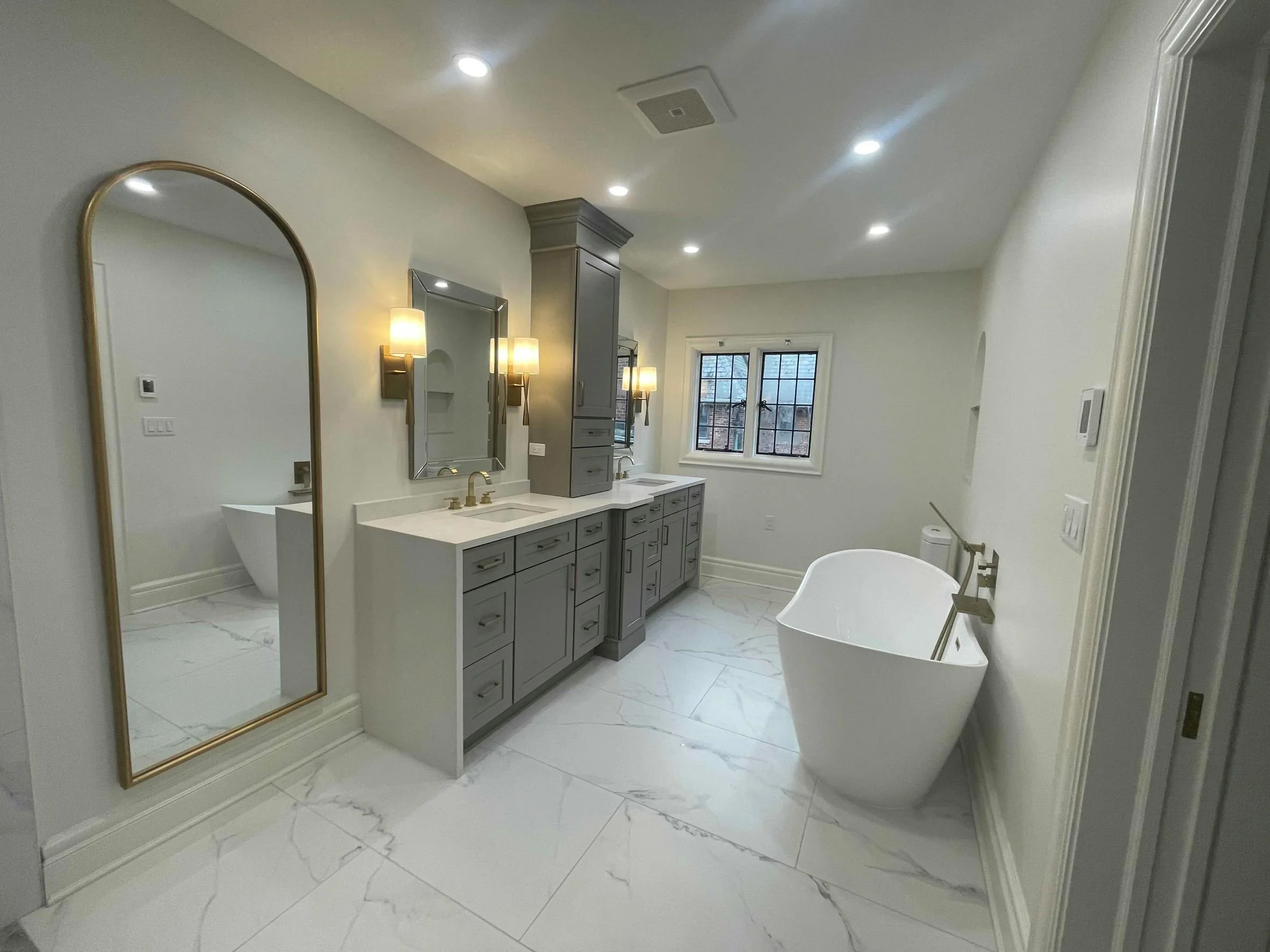 Modern bathroom featuring a freestanding bathtub, gray cabinets, a white countertop with double sinks, a large mirror, wall sconces, and a window overlooking a brick building.