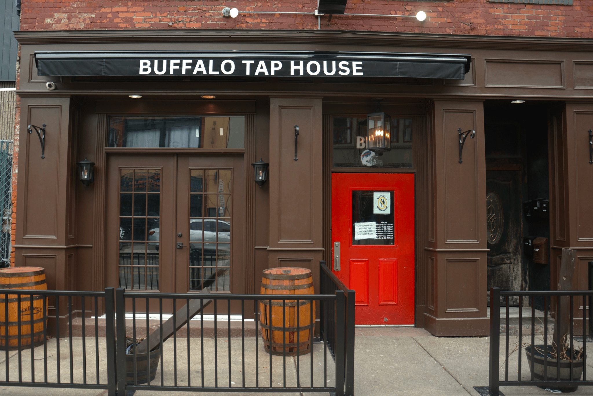 Exterior of a restaurant called Buffalo Tap House with a red door, brown brick and wood facade, barrel planters, and black fencing in front.