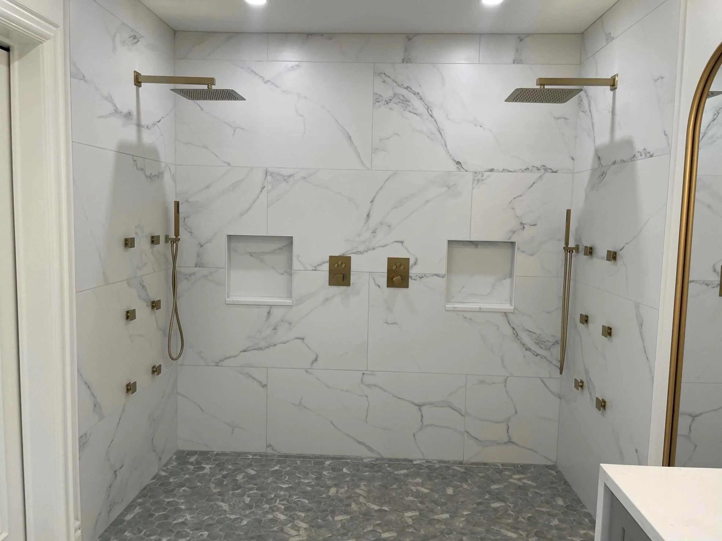 A modern walk-in shower with white marble walls, gold fixtures, two showers, and built-in niches.