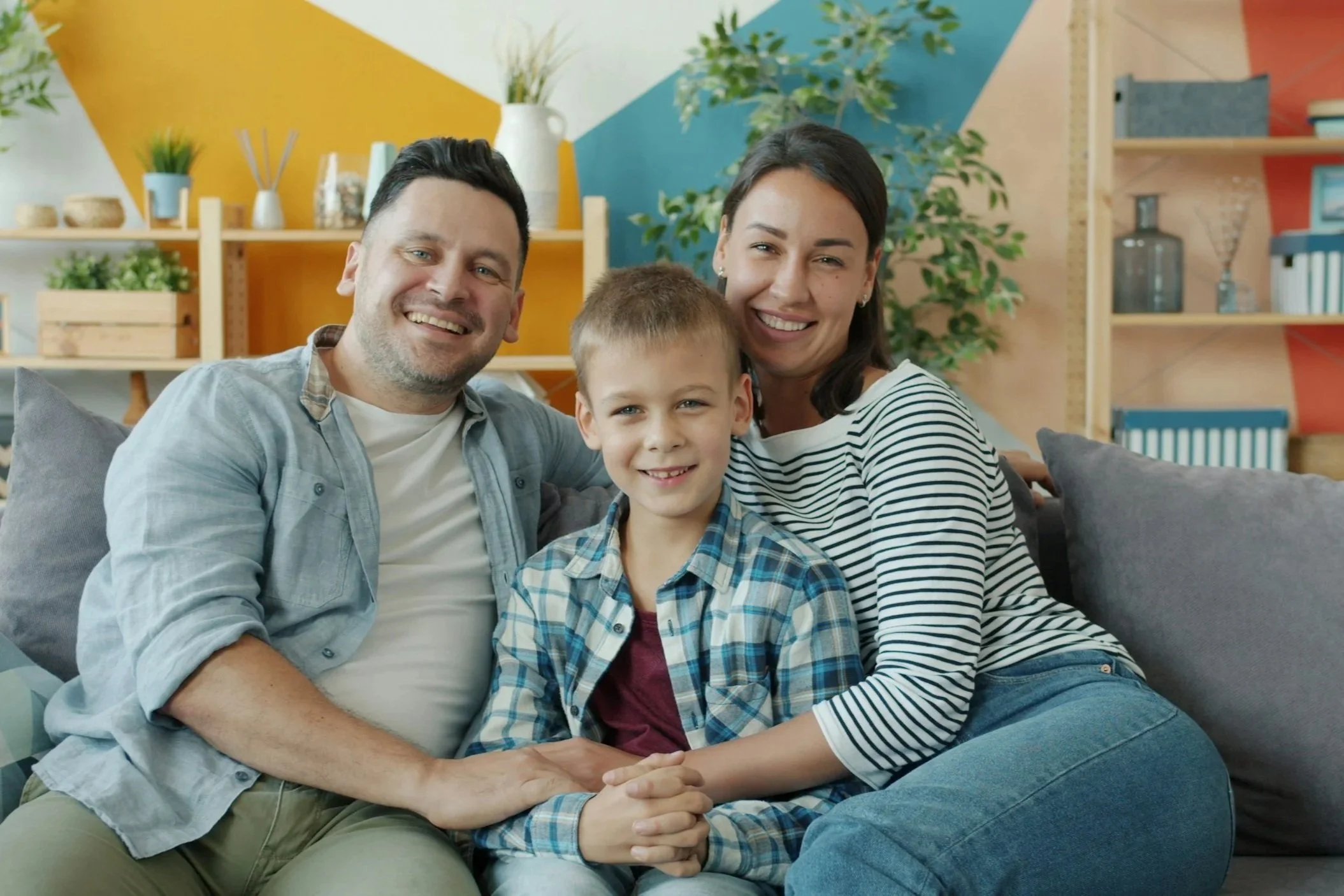 Family of three sitting closely on a couch, smiling at the camera, with colorful wall art and houseplants in the background.