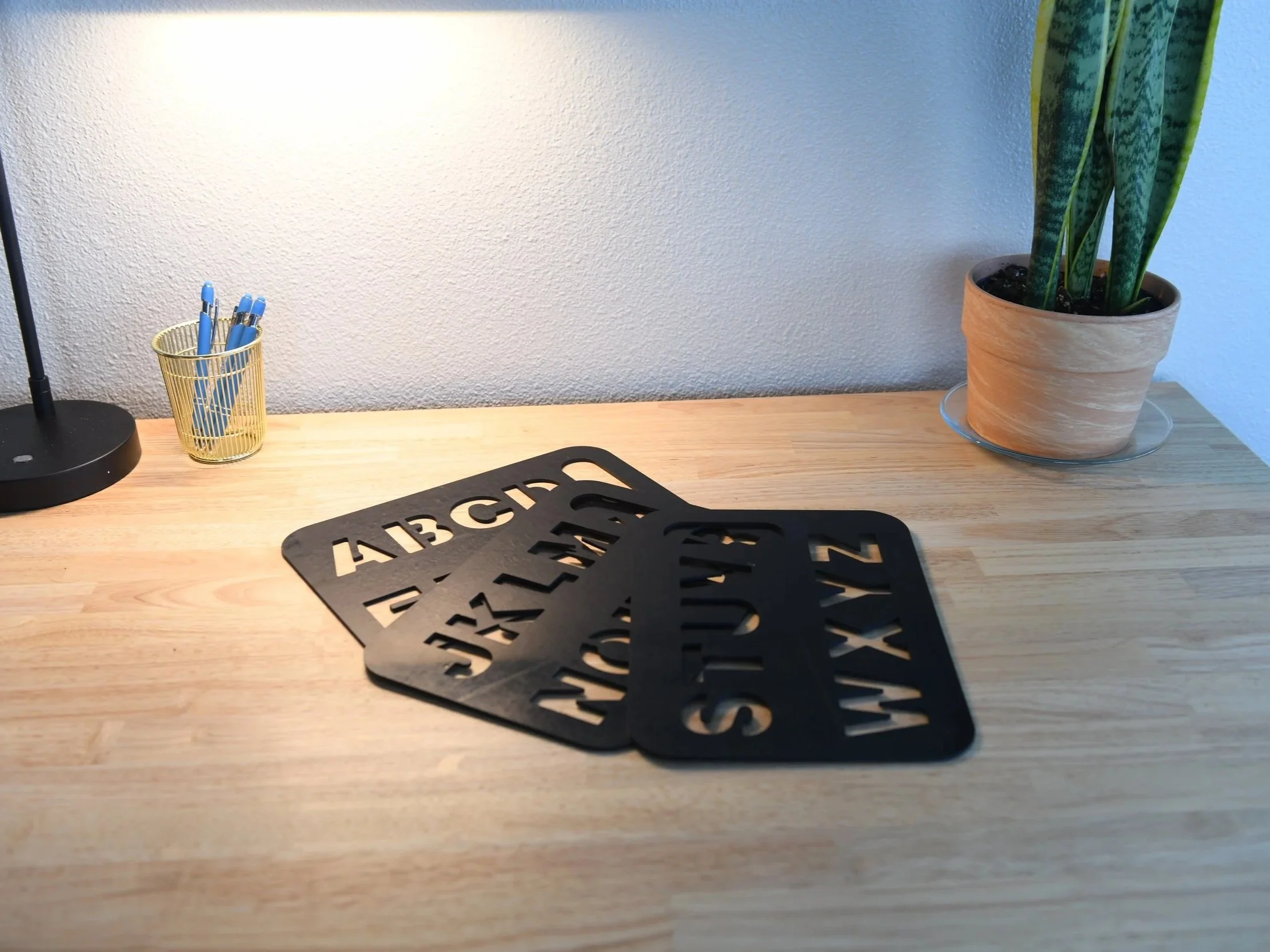 A wooden desk with a black lamp, a yellow pen holder with blue pens, three black stencils with letters, a potted snake plant in a beige pot, and a white wall in the background.
