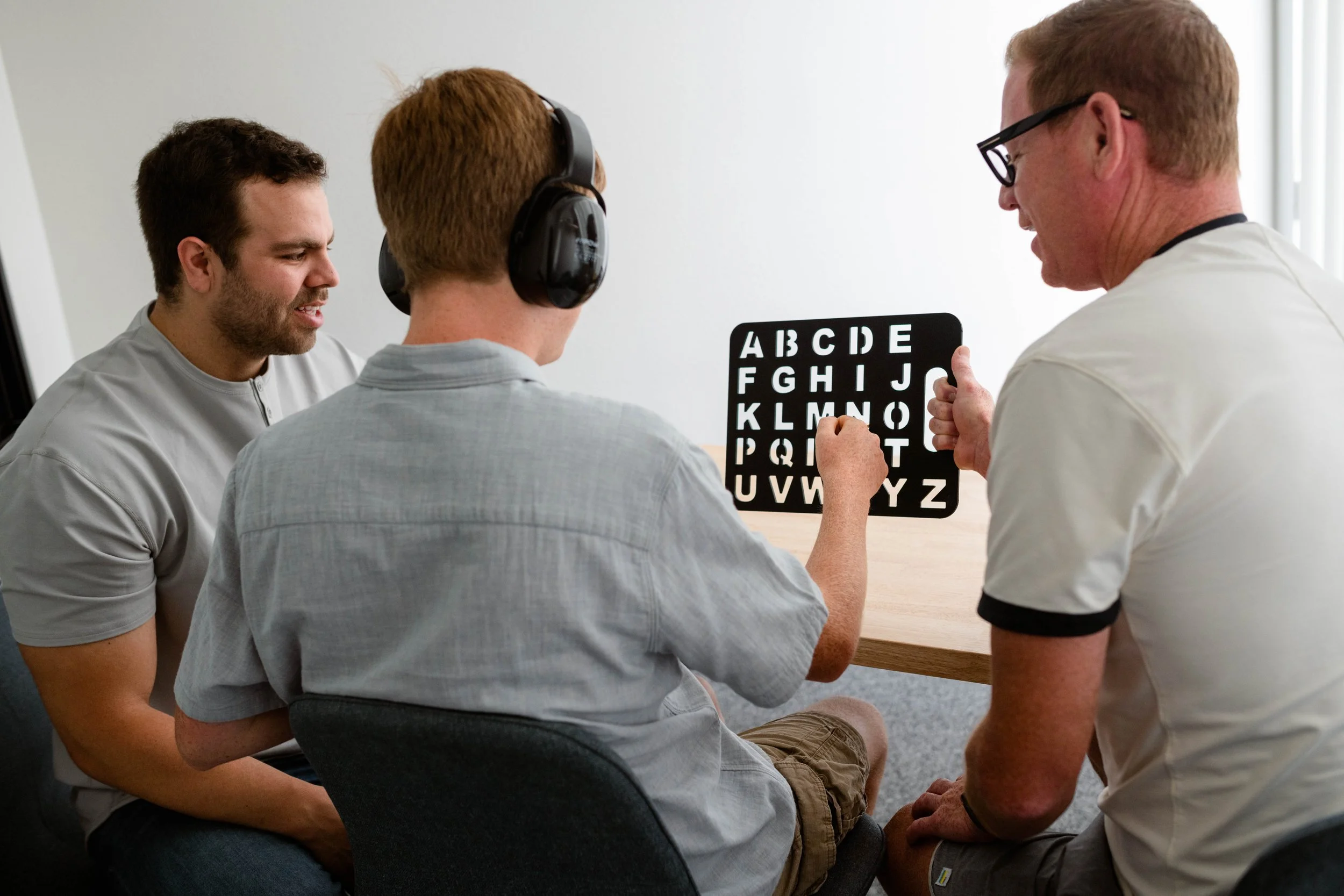 A speller wearing headphones is seated at a table pointing to letters on a stencil board. His father is seated on his right, holding the board encouragingly. Another man, the Spellers Method provider, is seated on the left coaching the parent.