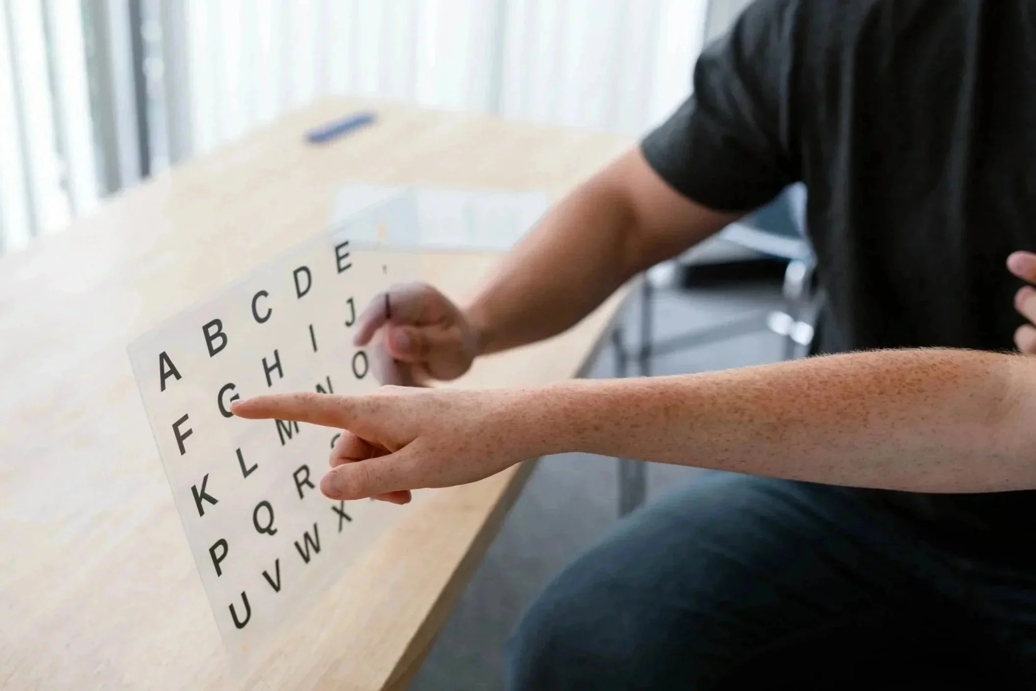 A speller pointing to the F on a laminated letterboard, seated at a light-colored table.