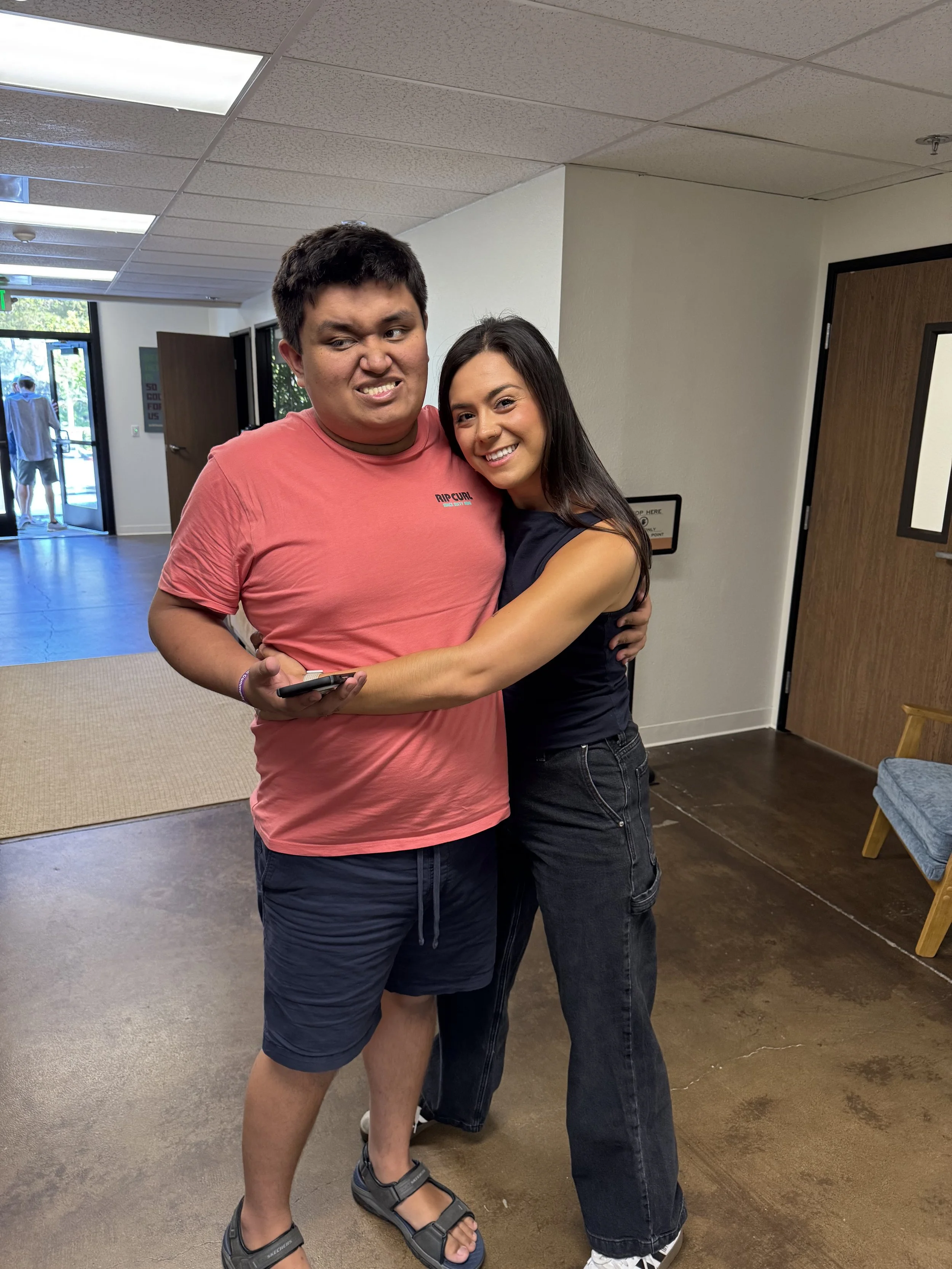 A young man and woman are hugging and smiling at the camera inside a building, with an open door showing people outside.