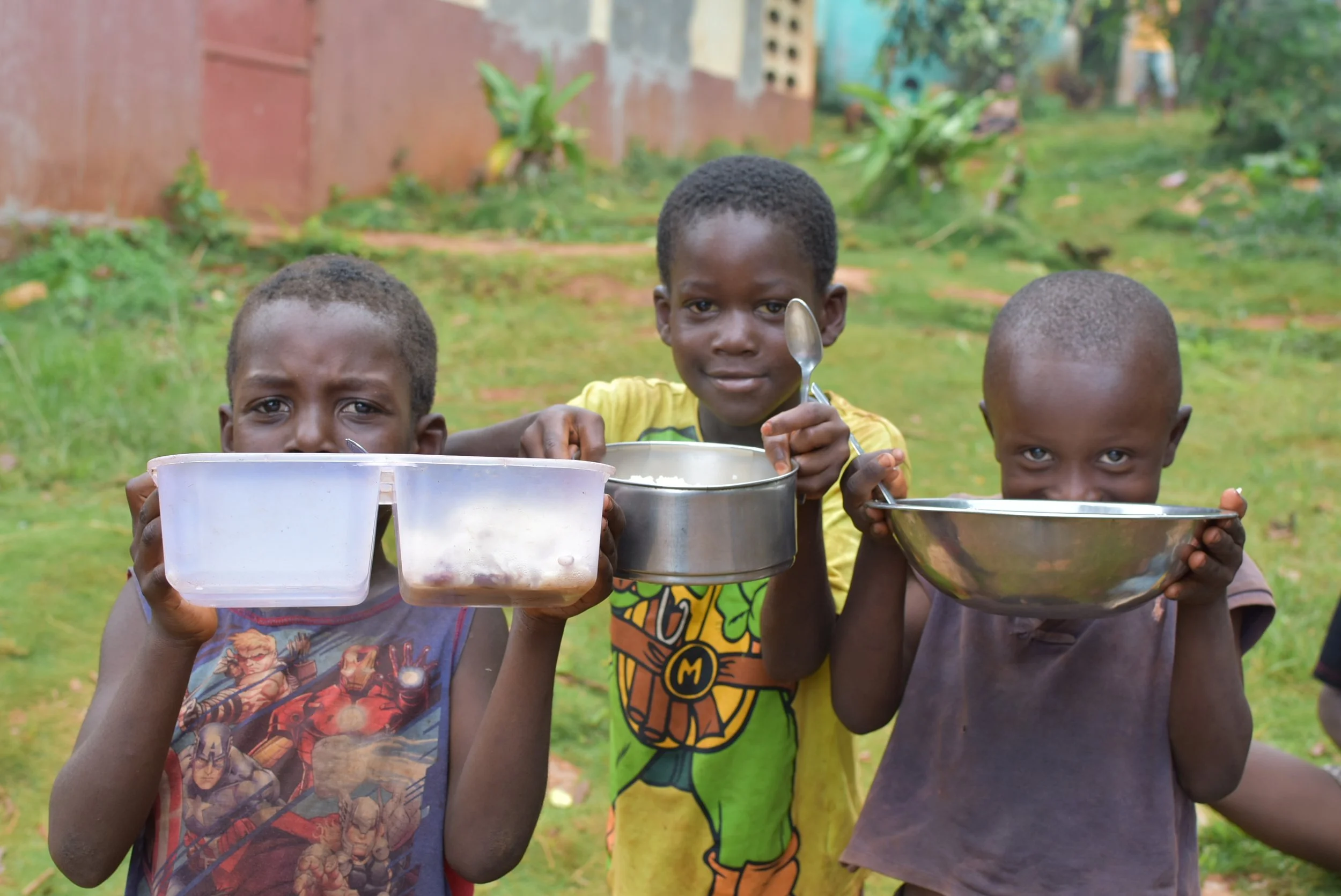 Three young boys holding containers with food outdoors, grassy area with plants and a brick wall in the background.