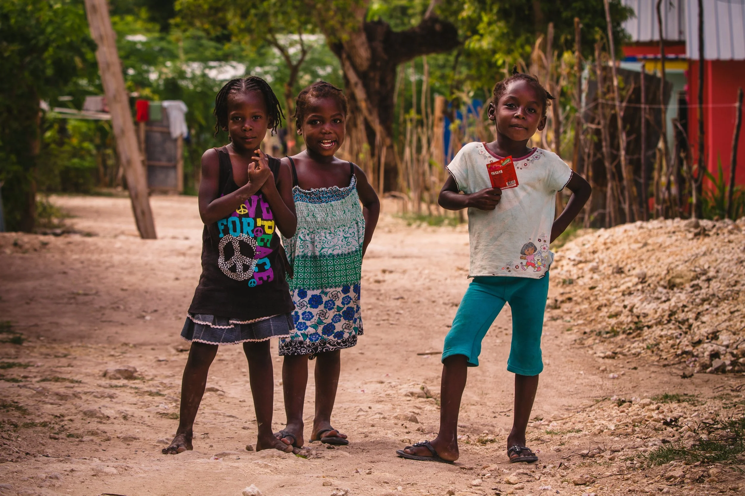 Three young girls standing on a dirt path in a rural area with trees and simple houses in the background.