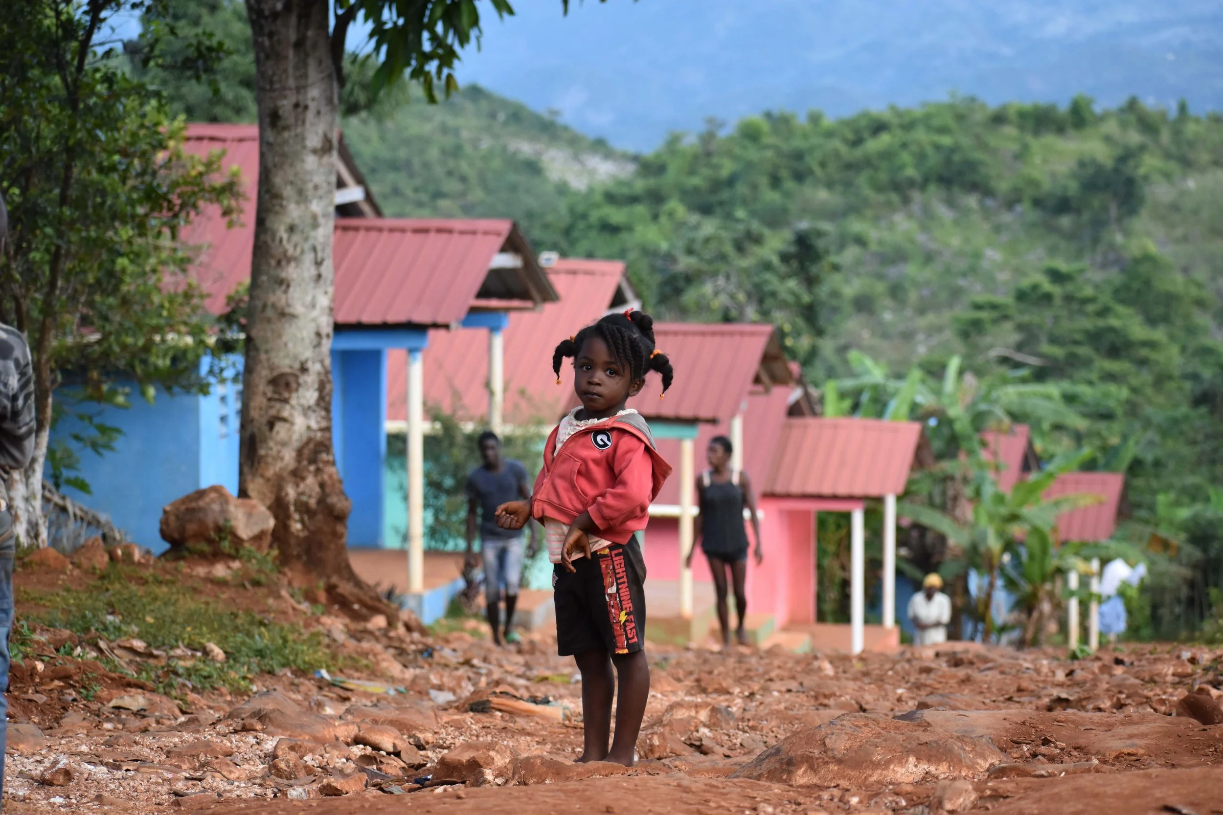 A young girl with pigtails standing on a rocky dirt path in a rural area, with colorful houses, trees, and mountains in the background.