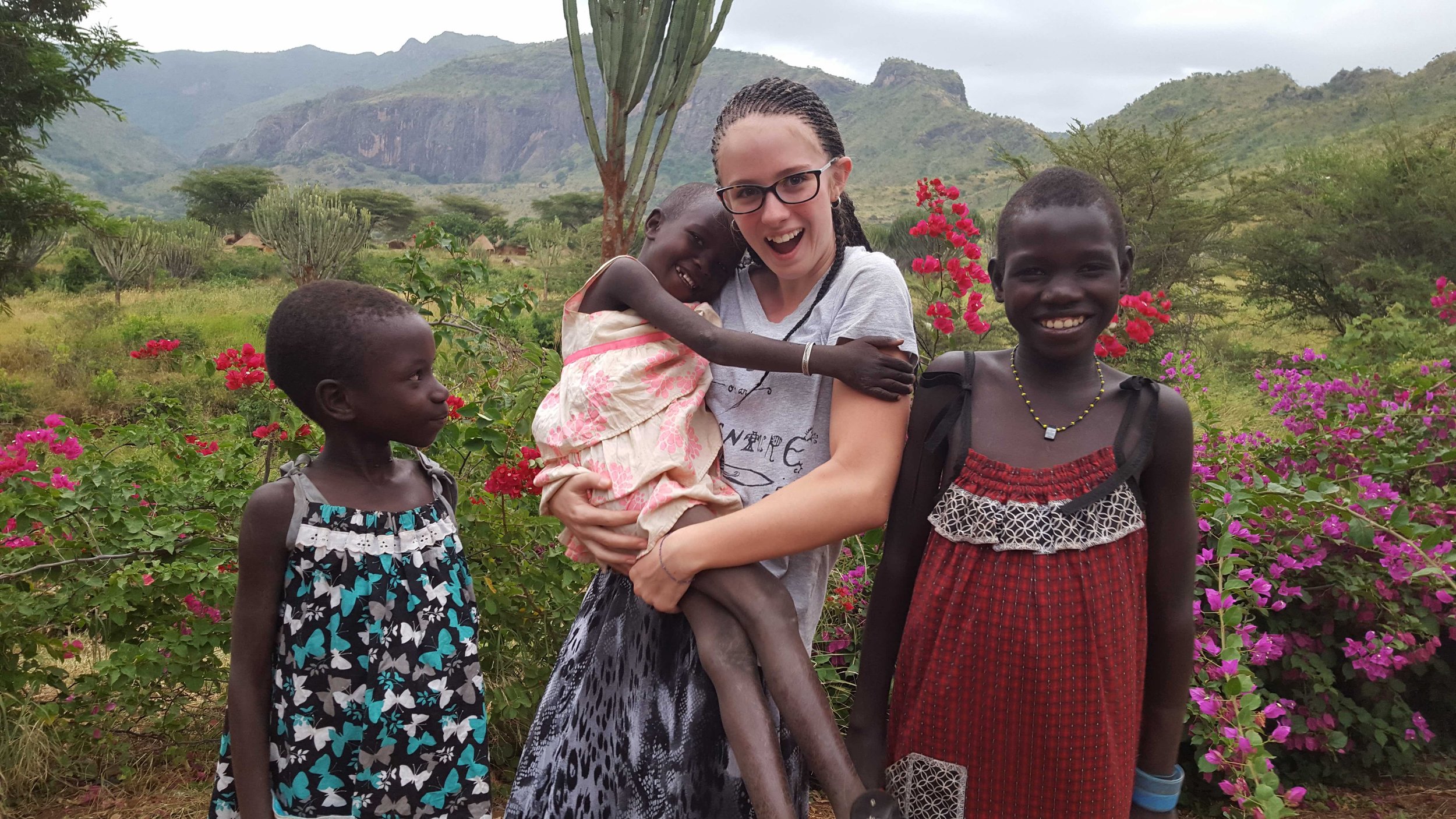 A woman and three young girls standing outdoors amid flowering bushes and mountainous terrain, smiling and posing for the photograph.