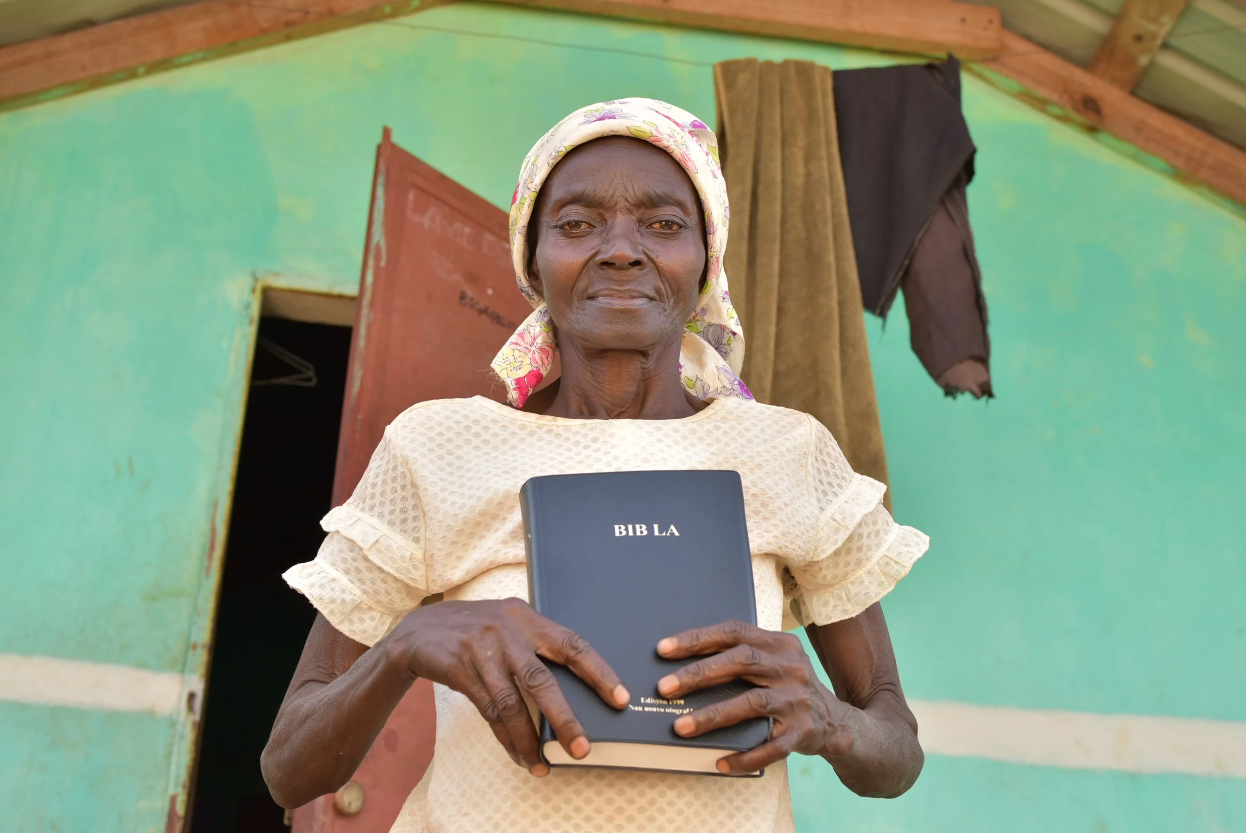 An elderly woman stands outside a green dwelling, holding a Bible with the title 'BIB LA', wearing a patterned headscarf and a beige blouse.