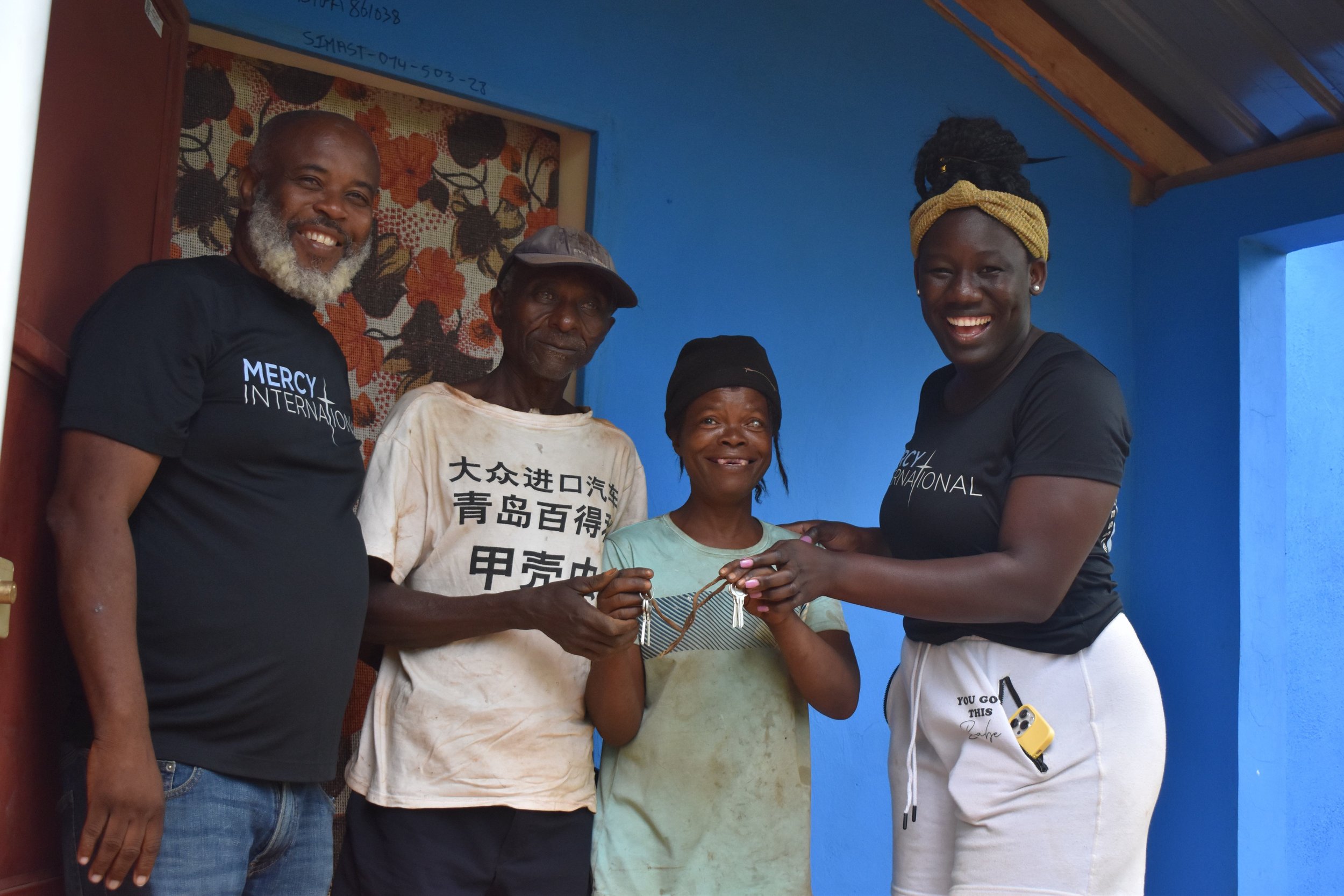 Four people, two women and two men, standing outside a blue building, smiling, with one woman holding a necklace, two people wearing Mercy International t-shirts.
