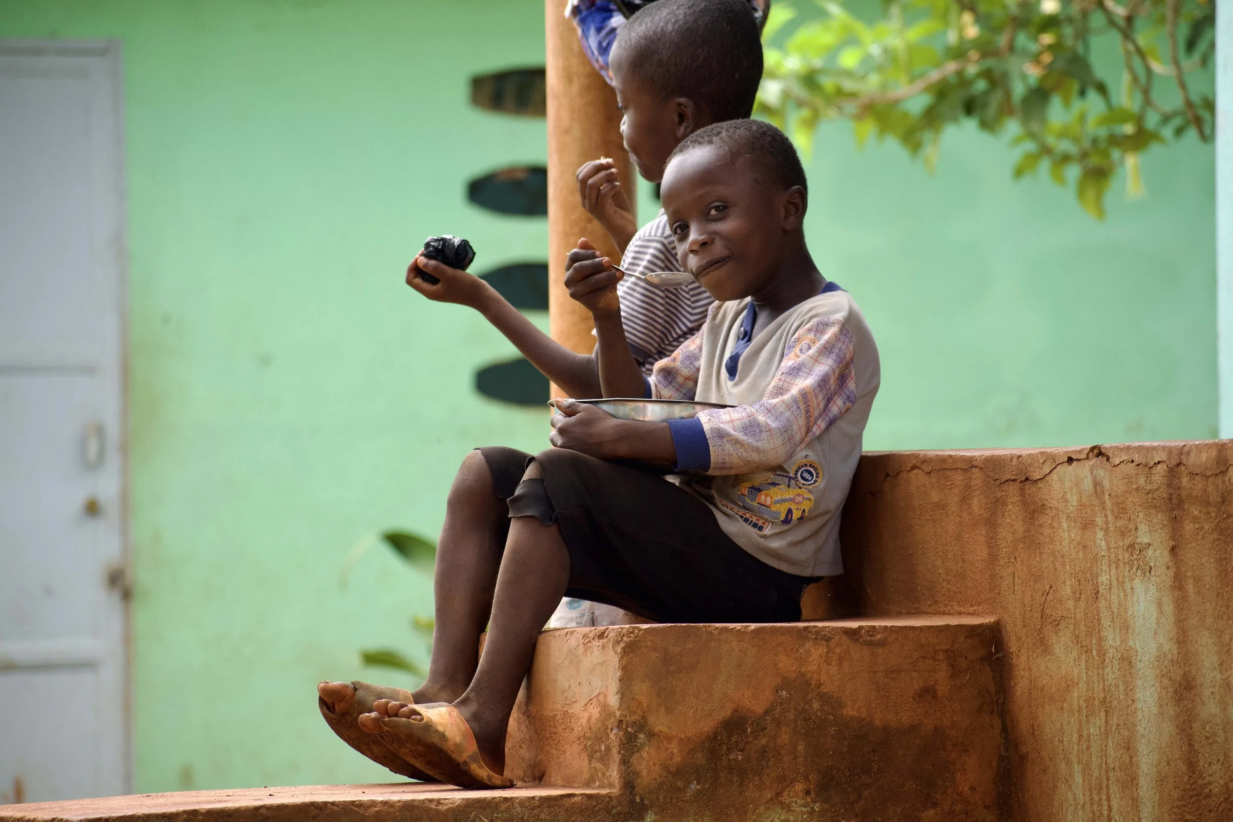 Two young boys sitting on an orange concrete ledge, one eating with a spoon and the other holding a black object, against a green wall with leaves.