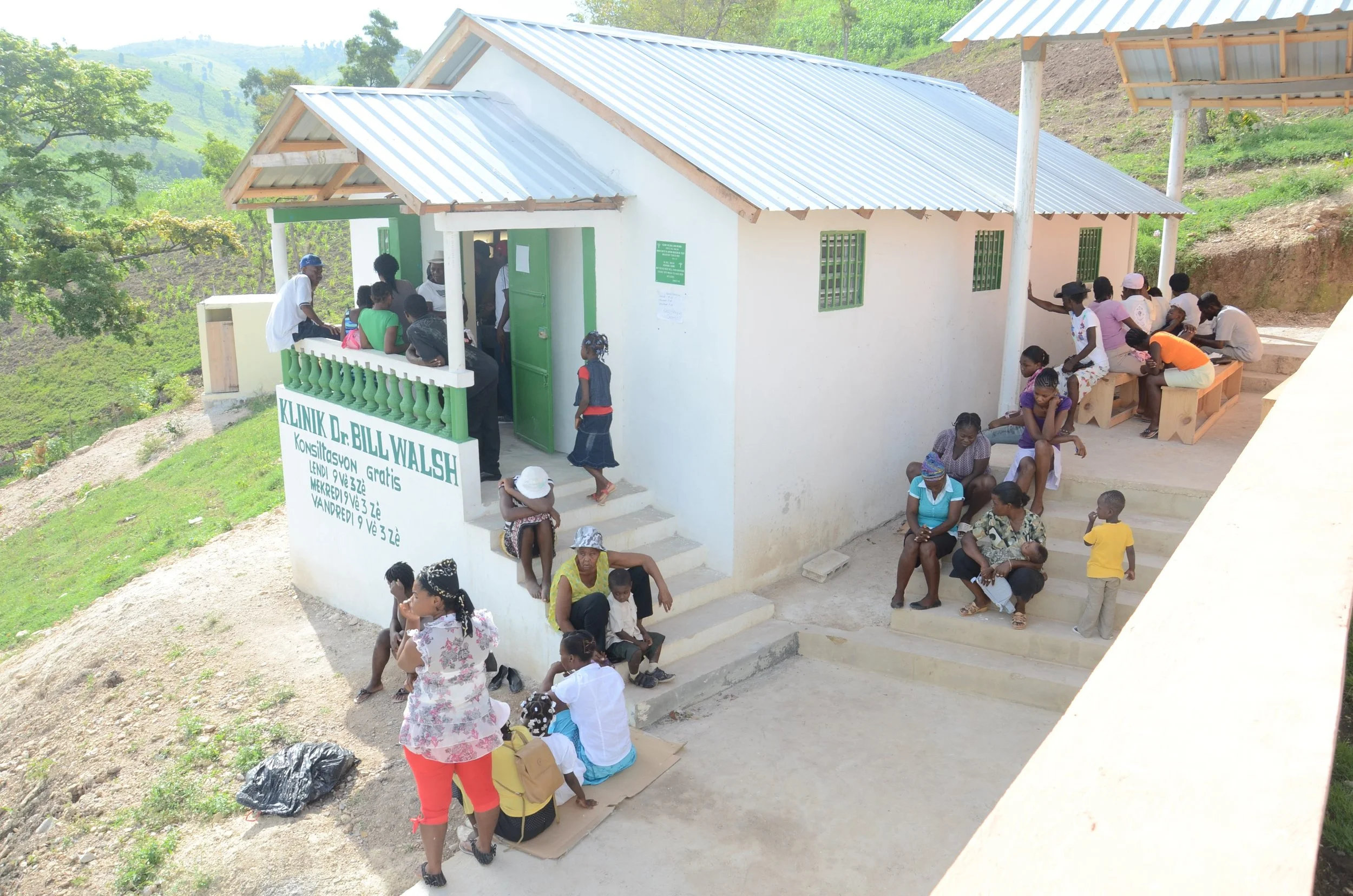 A small clinic building with a green roof and white walls, with many people waiting outside on a sunny day in a rural area with green hills in the background.